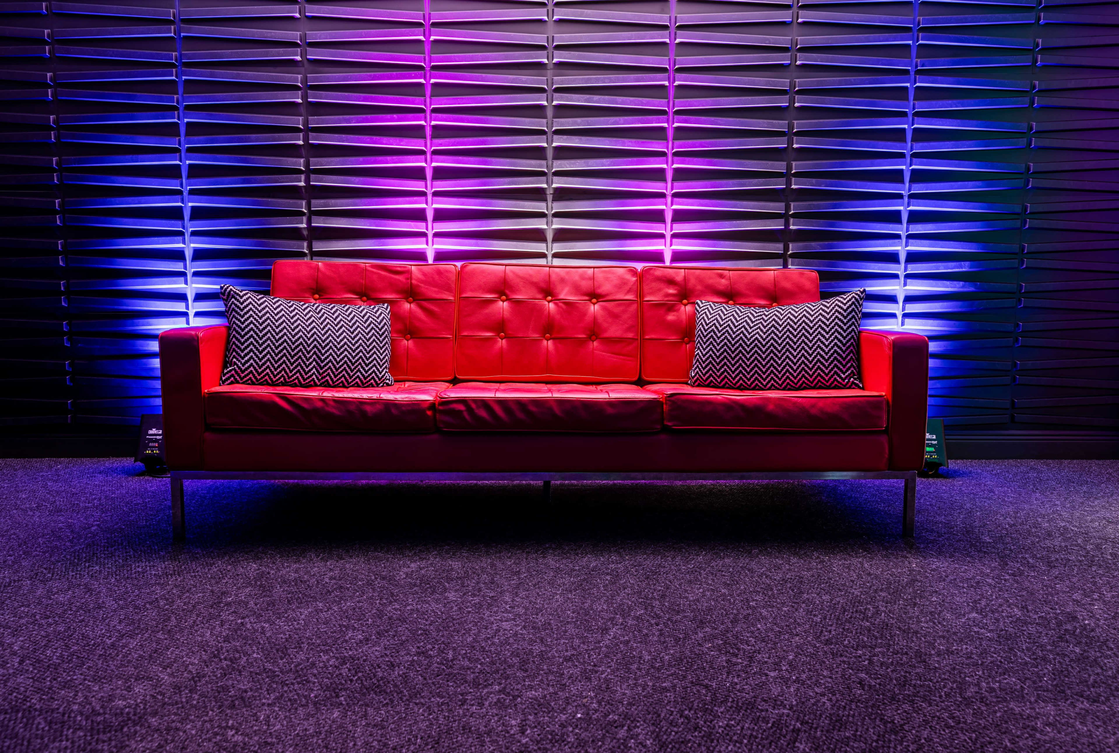 A red sofa with two patterned cushions is positioned against a textured wall illuminated in purple and blue light.