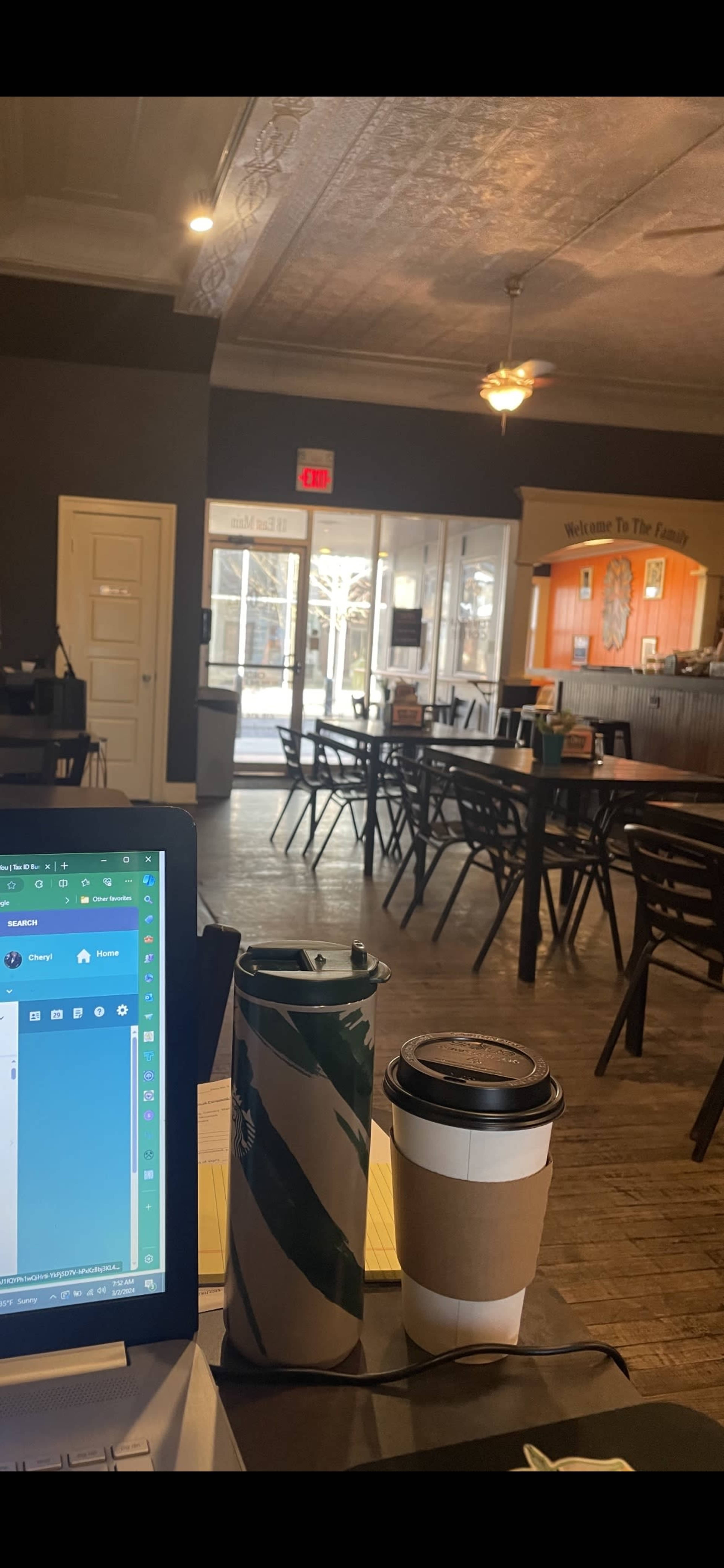 The image shows a quiet café interior with several empty tables, a laptop on a table, and two coffee cups in the foreground.