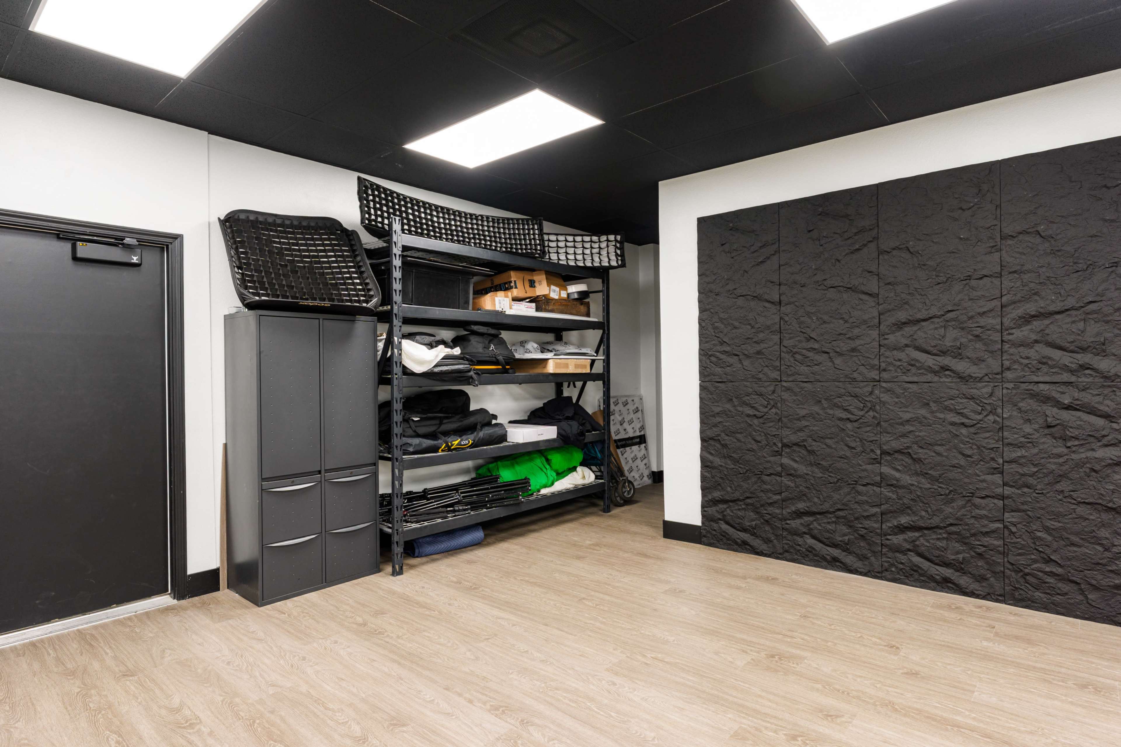 The image shows a storage room with shelving units filled with various items and a black wall panel.
