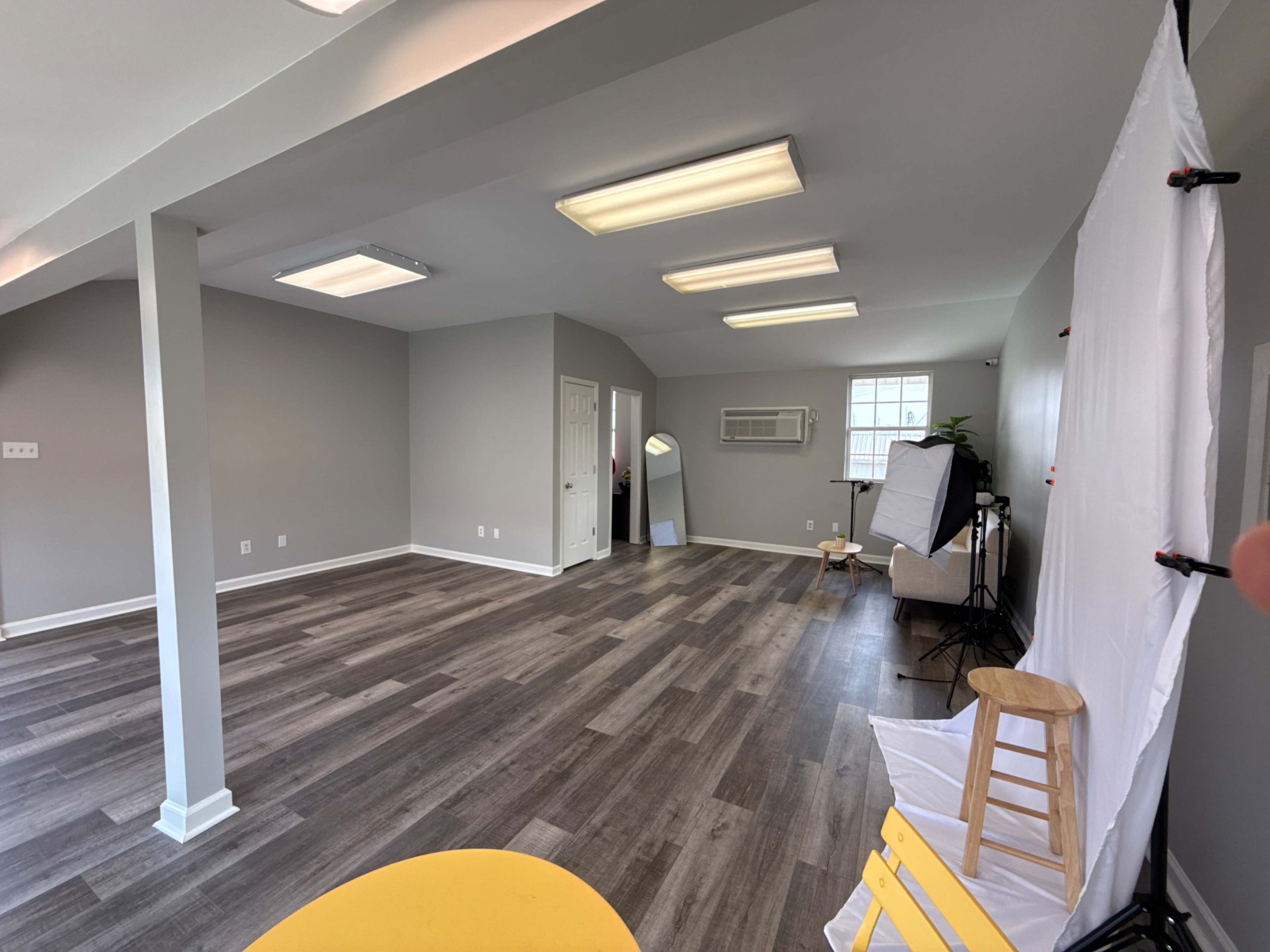 The image shows a spacious, well-lit room with gray walls, wooden flooring, and minimal furniture, including a few stools and a backdrop setup for photography.