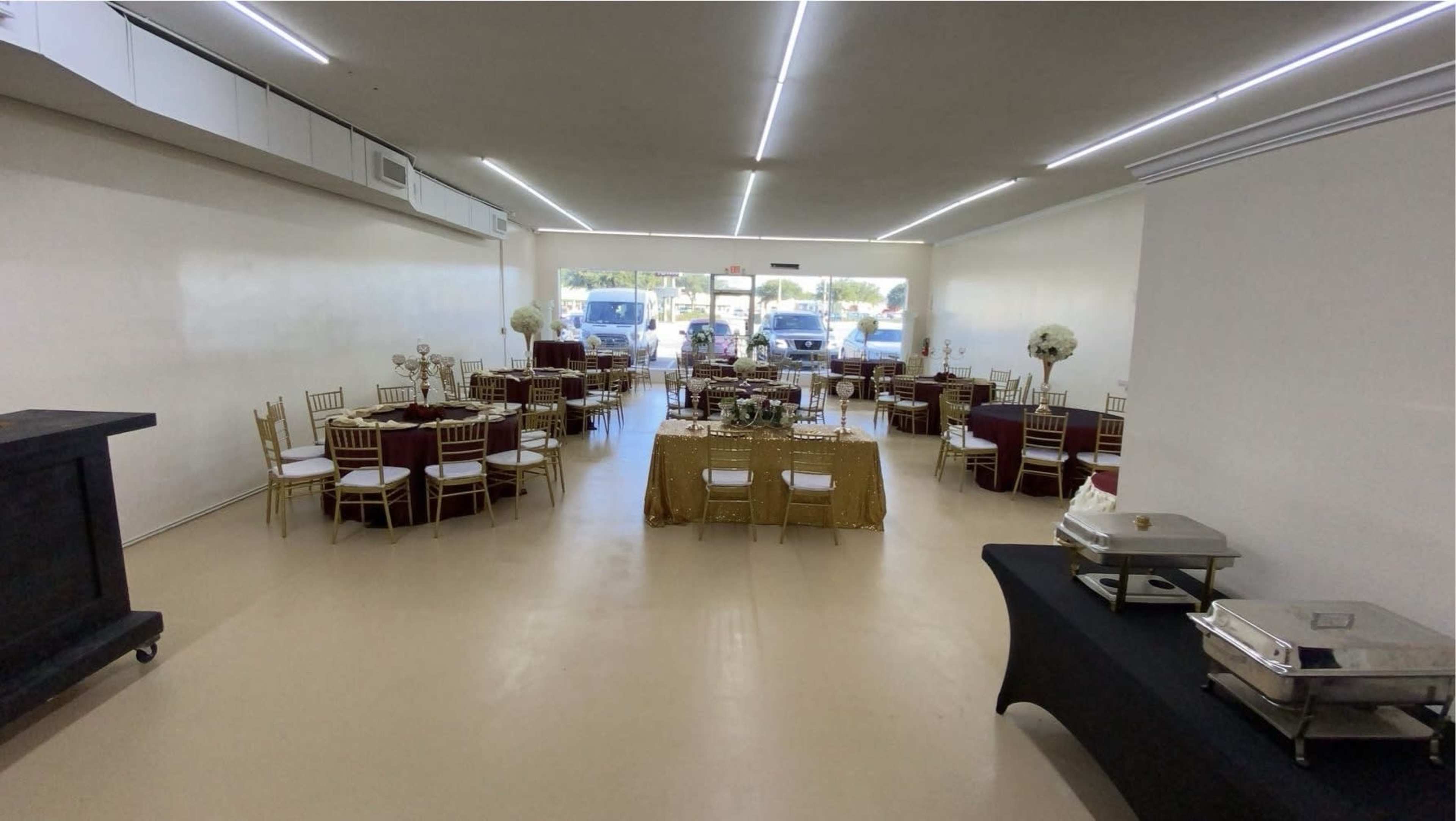 A spacious event venue set up with tables covered in various colored linens, gold-accented decor, and chafing dishes on a side table, with large windows allowing natural light to enter.