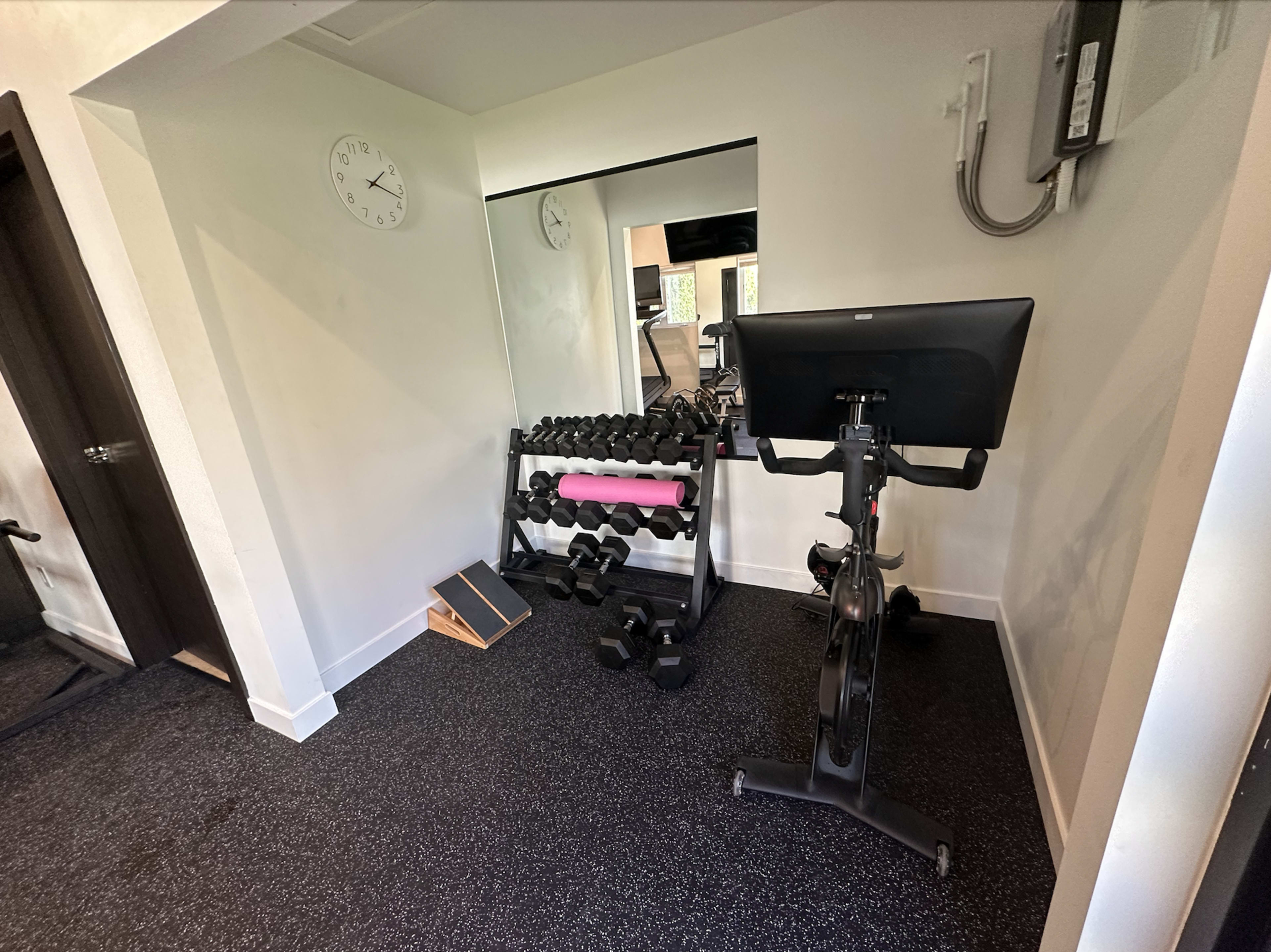 The image shows a small home gym featuring a rack of dumbbells, a stationary bike, and a large wall mirror.