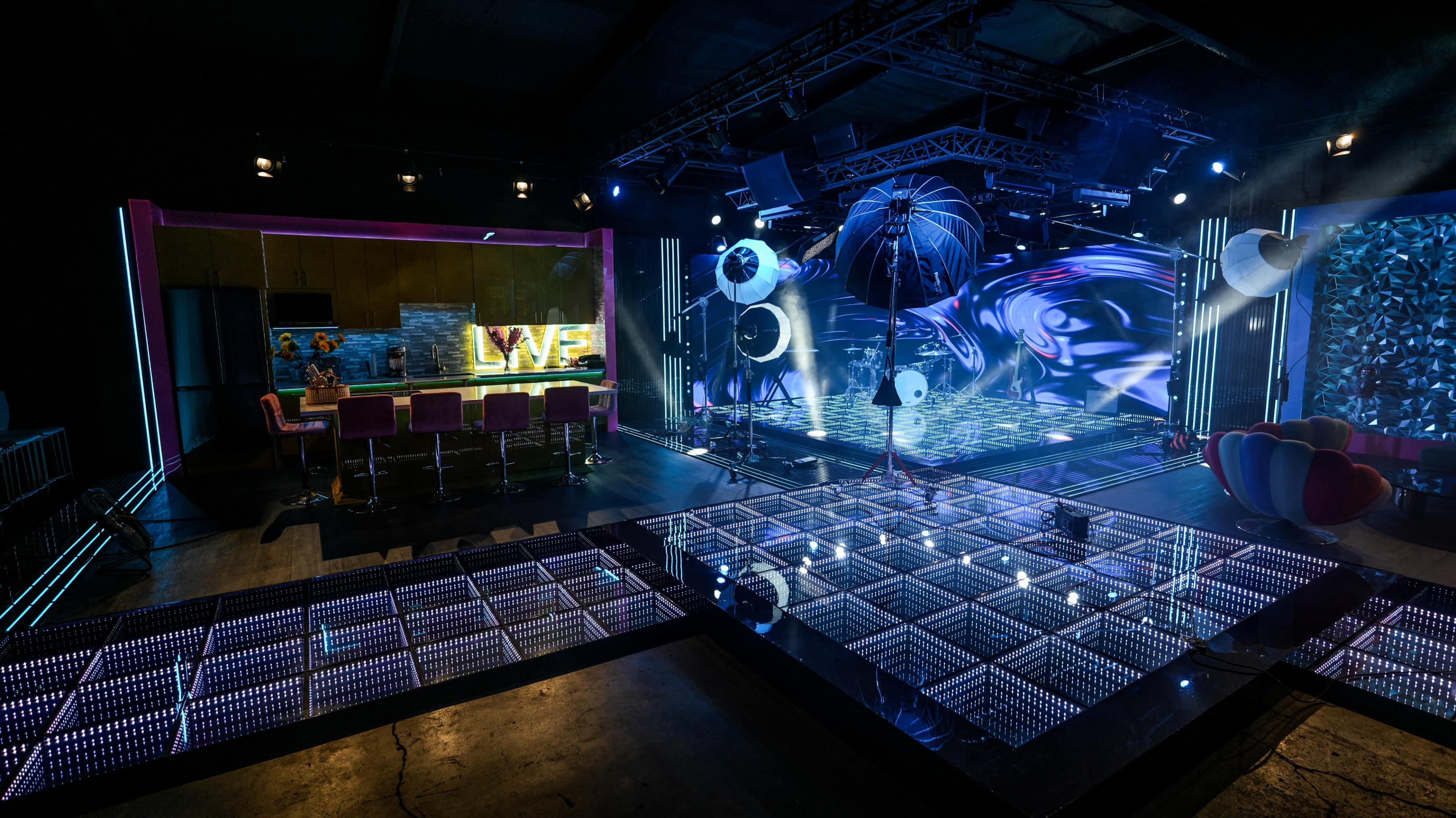 The image shows a modern television studio set with colorful lighting, a bar area, and a large digital backdrop displaying abstract visuals.