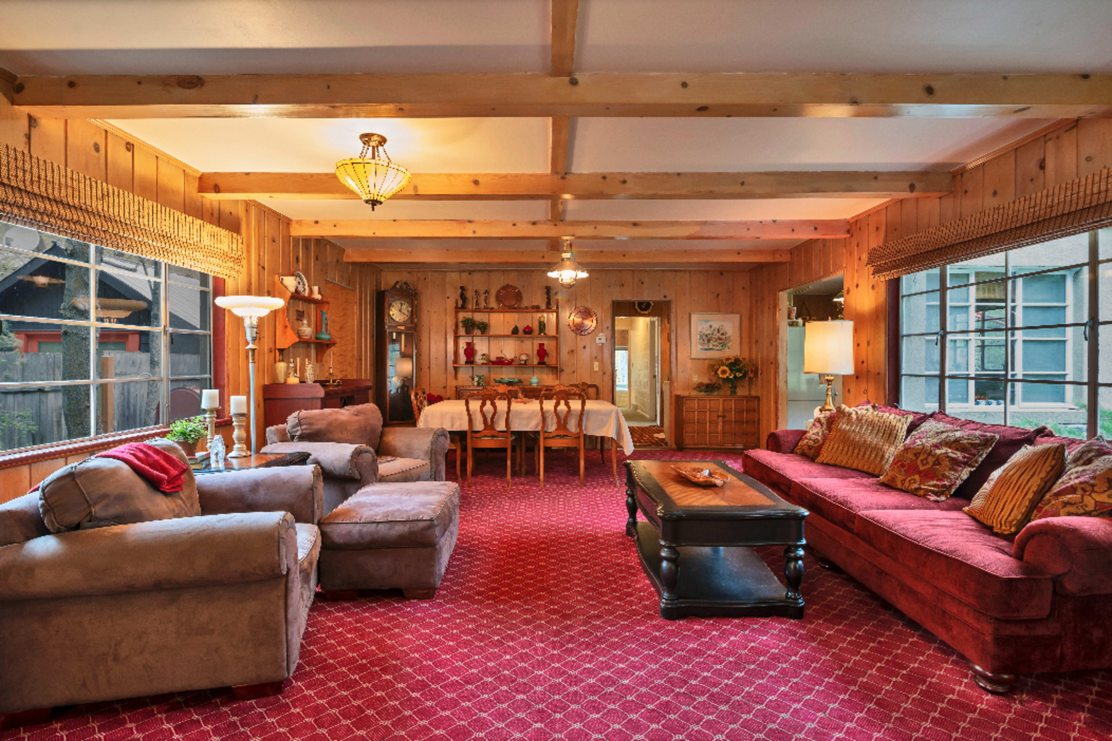 A cozy living room with wooden walls, a red carpet, a dining area in the background, and comfortable seating arrangements including a sofa and armchairs.