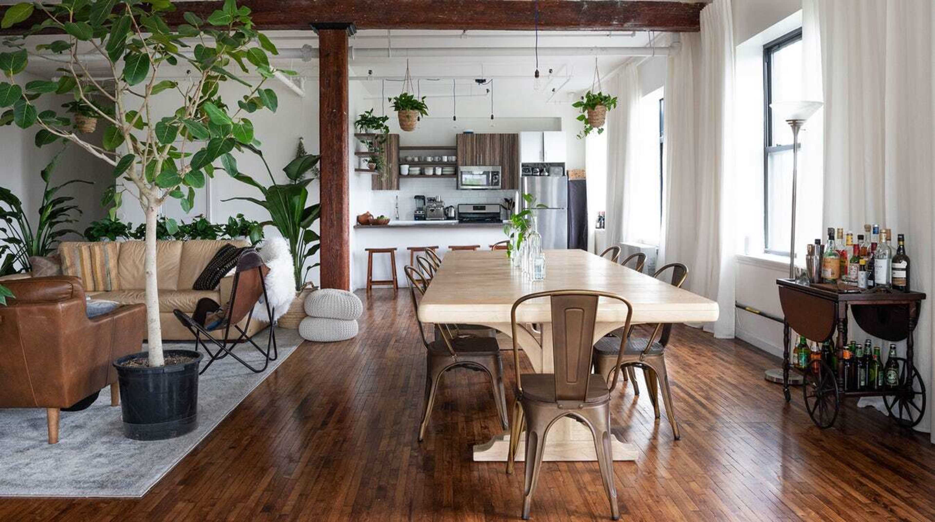 A modern, open-concept living and dining area with a wooden dining table surrounded by metal chairs, a sofa, and plants, all set against a backdrop of large windows and wooden beams.