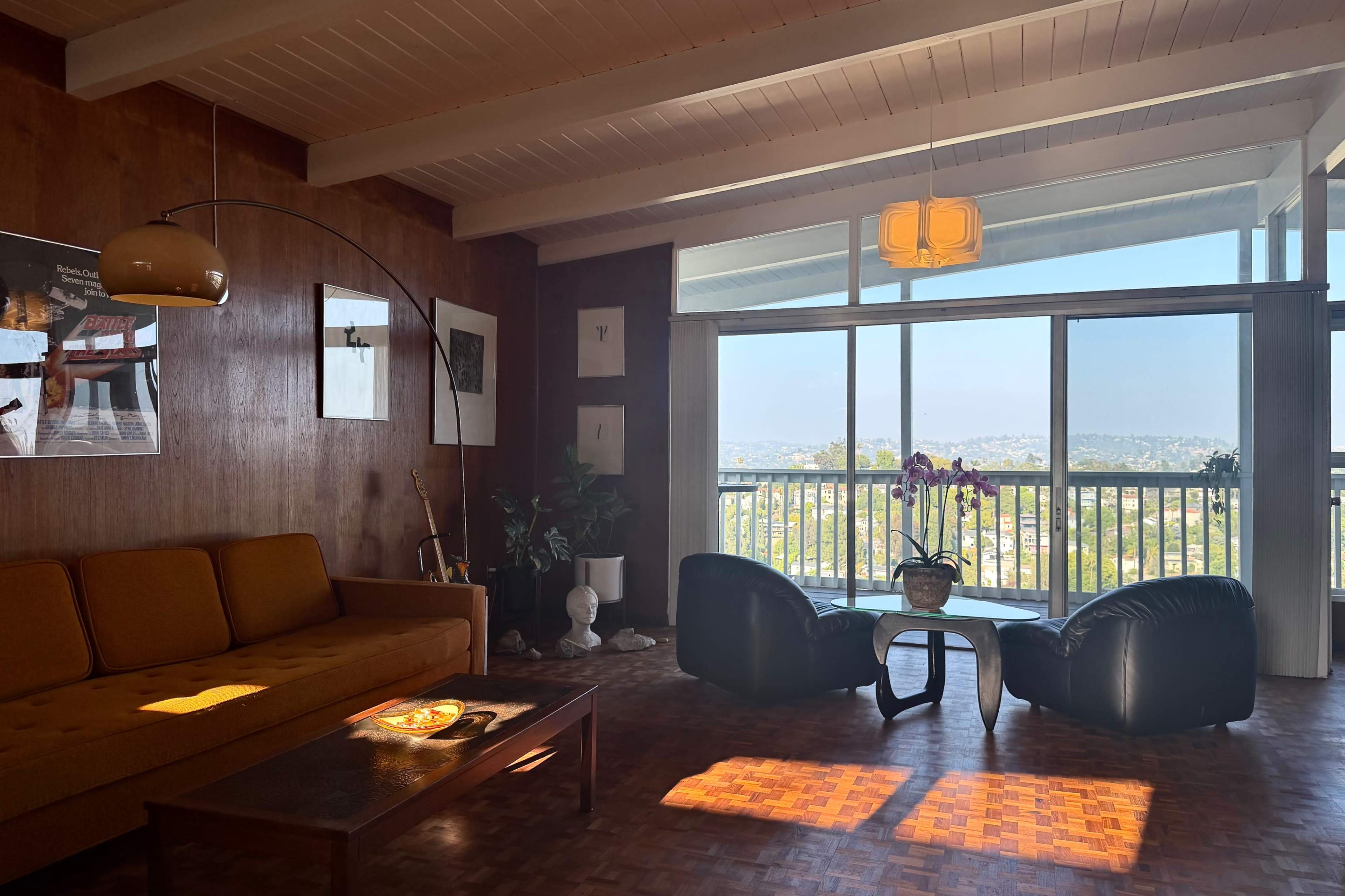 The image shows a mid-century modern living room with a brown sofa, a coffee table, and large windows that offer a view of the landscape outside.