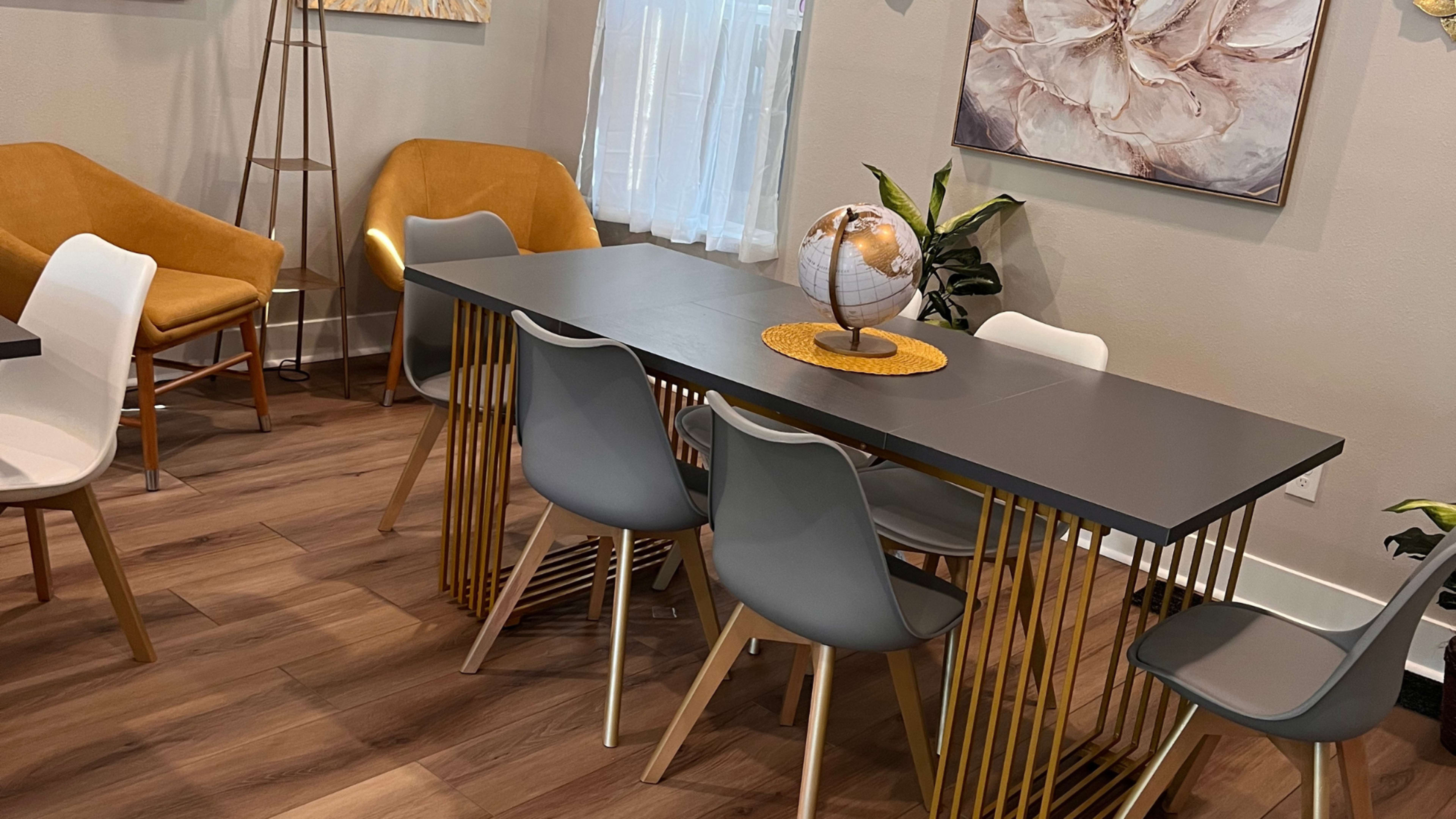 A modern dining area features a table with a gray surface and gold legs surrounded by gray and white chairs, with two yellow armchairs in the background.