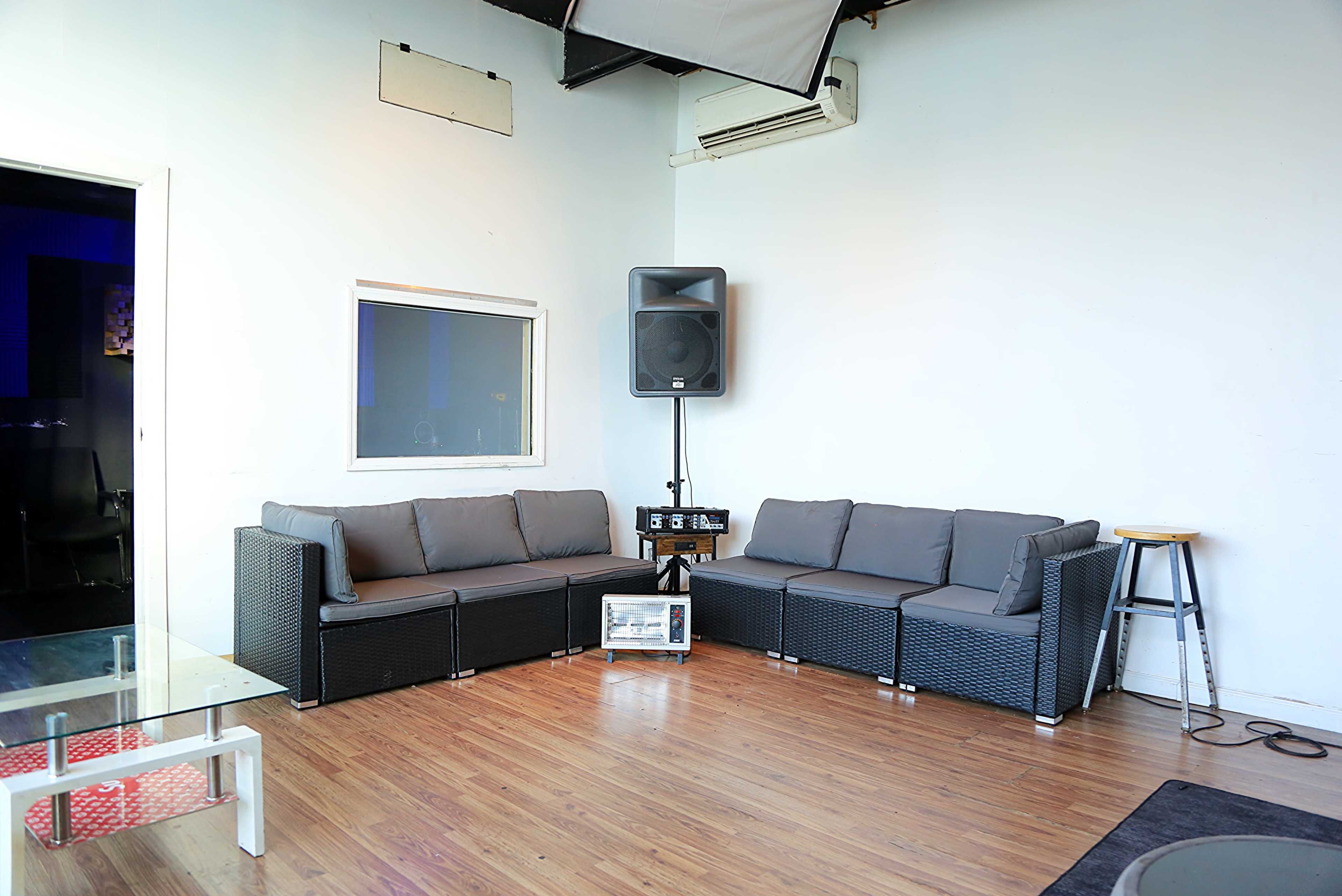 The image shows a minimalist indoor lounge area with a gray sectional couch, a small table, and a speaker mounted on the wall.