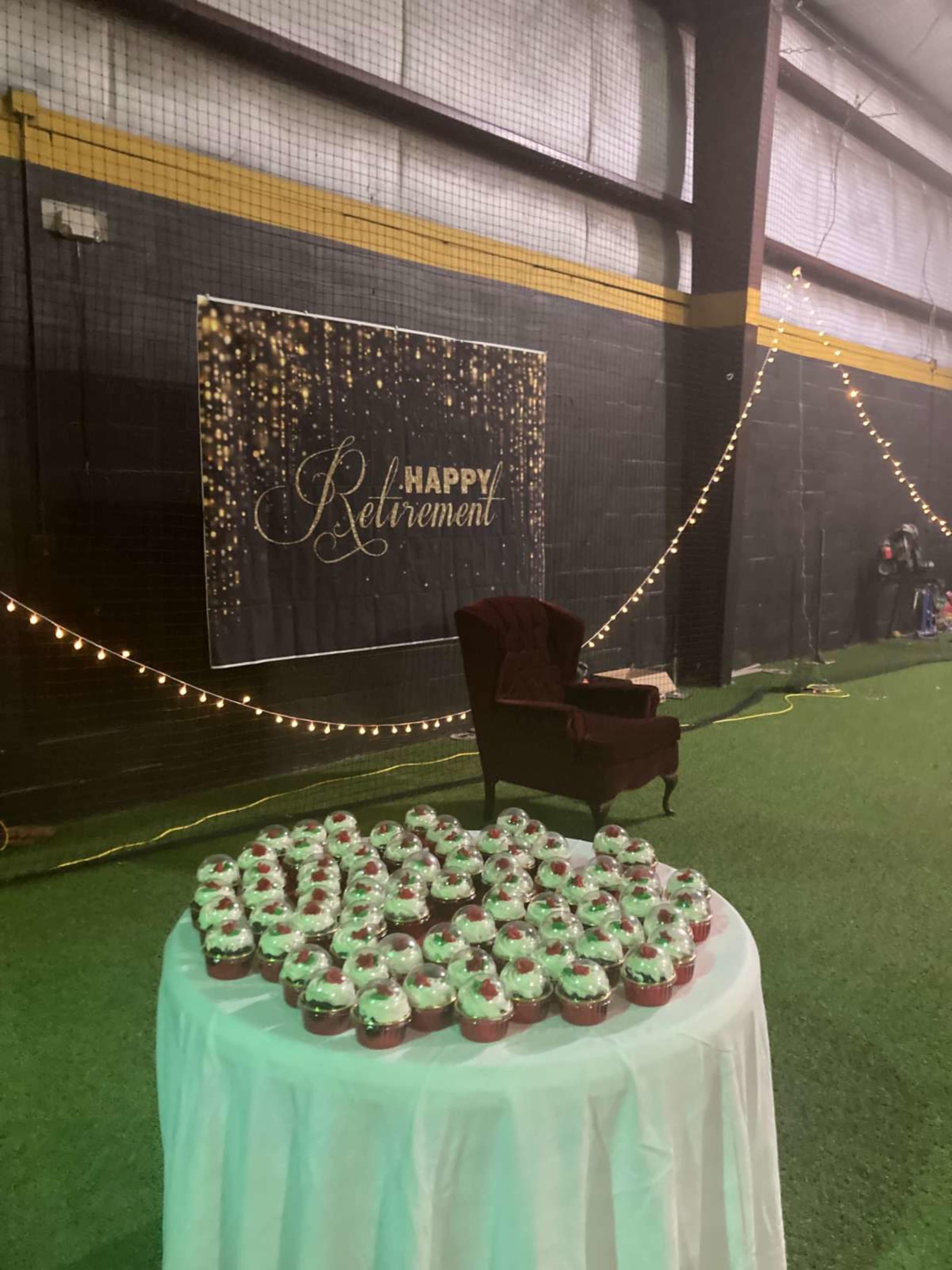 A table with cupcakes is set up in front of a "Happy Retirement" banner in a decorated indoor space with string lights.