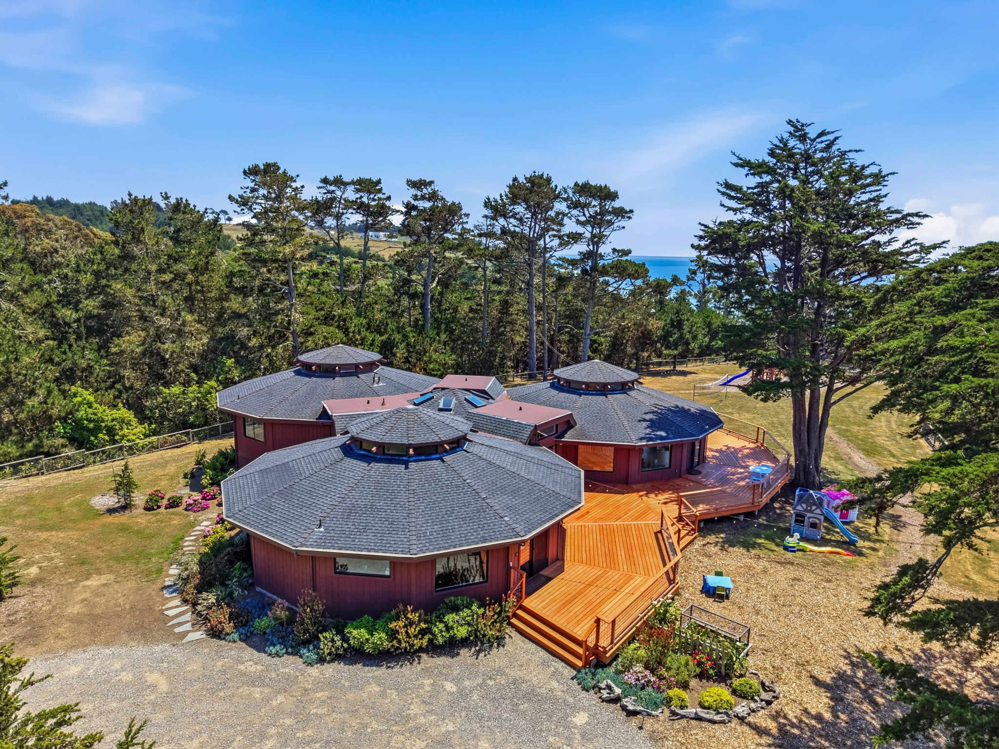 The image shows a large, circular house with multiple sections surrounded by trees and a grassy area, overlooking a body of water in the distance.