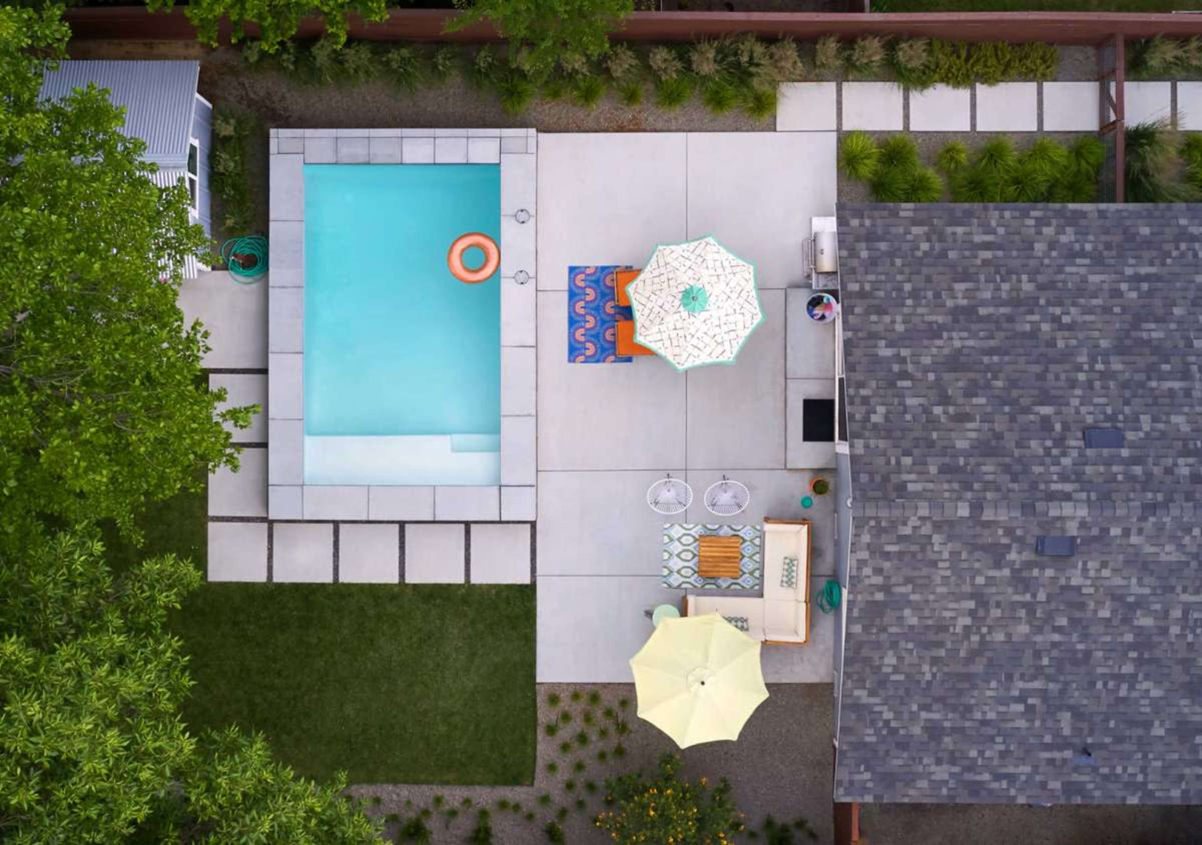 The image shows a backyard with a rectangular swimming pool surrounded by a patio area featuring lounge chairs, umbrellas, and a lawn.