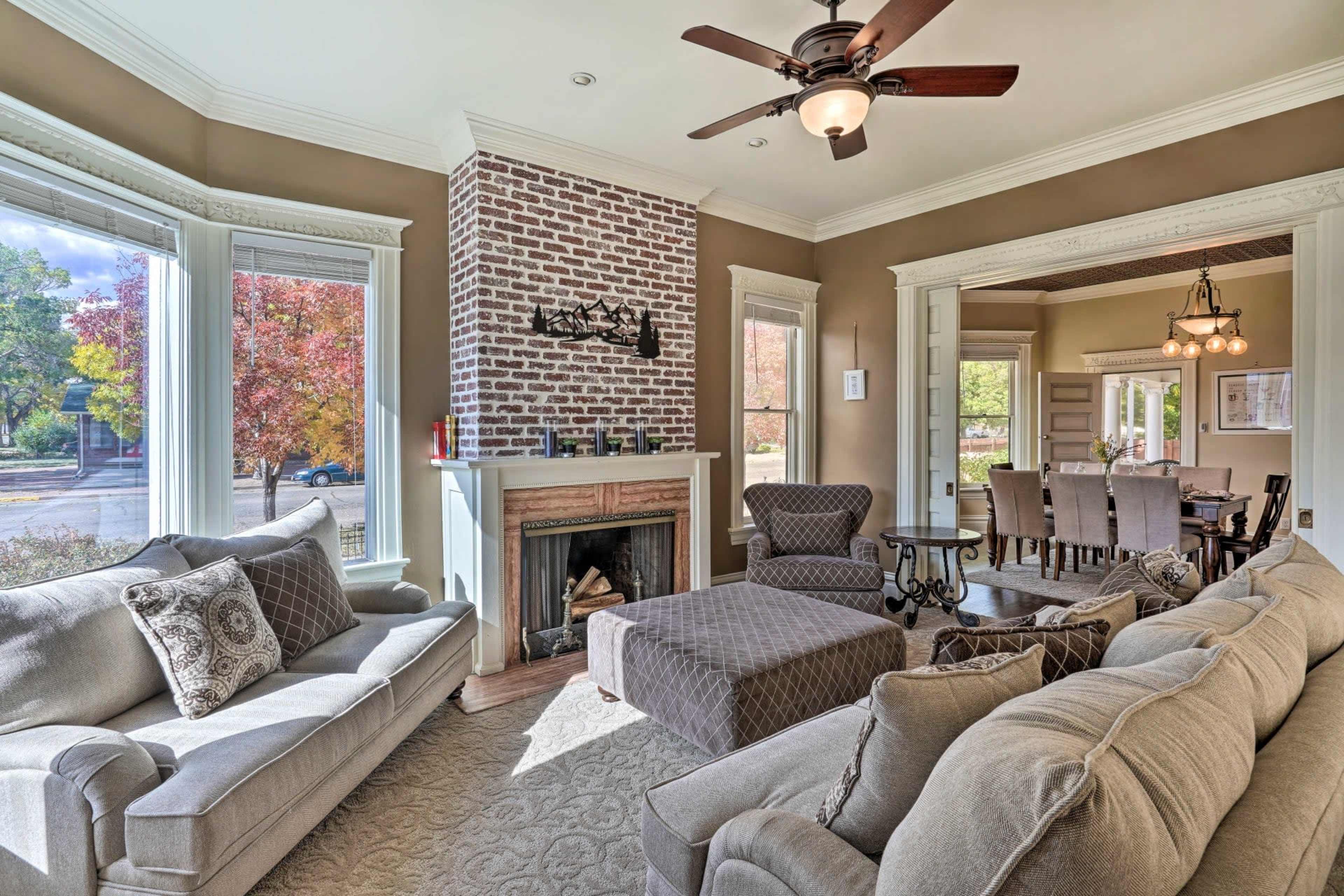 A cozy living room features a brick fireplace, large windows, and comfortable seating with a view of an outdoor scene.