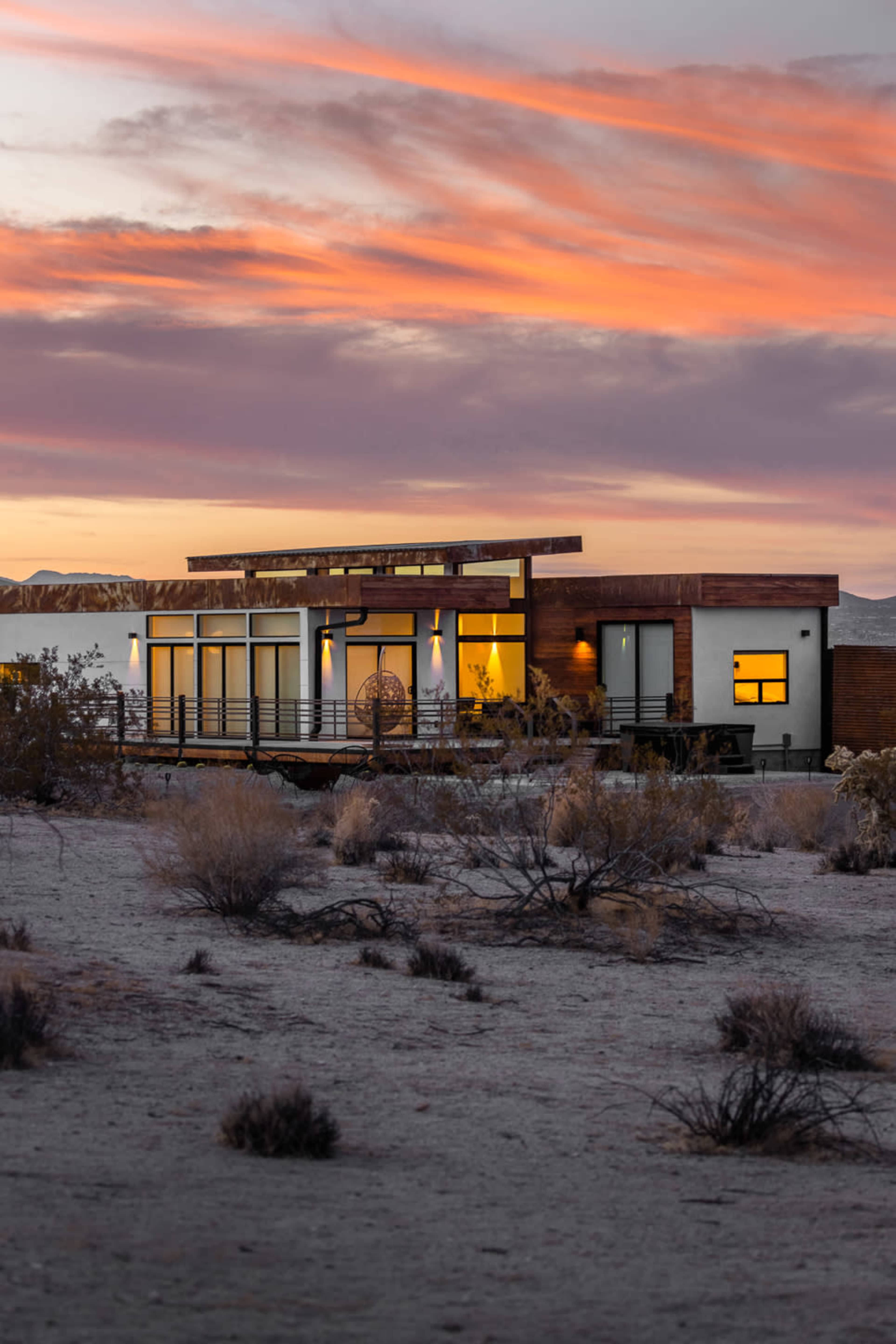 A modern house with large windows and a wooden exterior is set against a colorful sunset sky over a desert landscape.