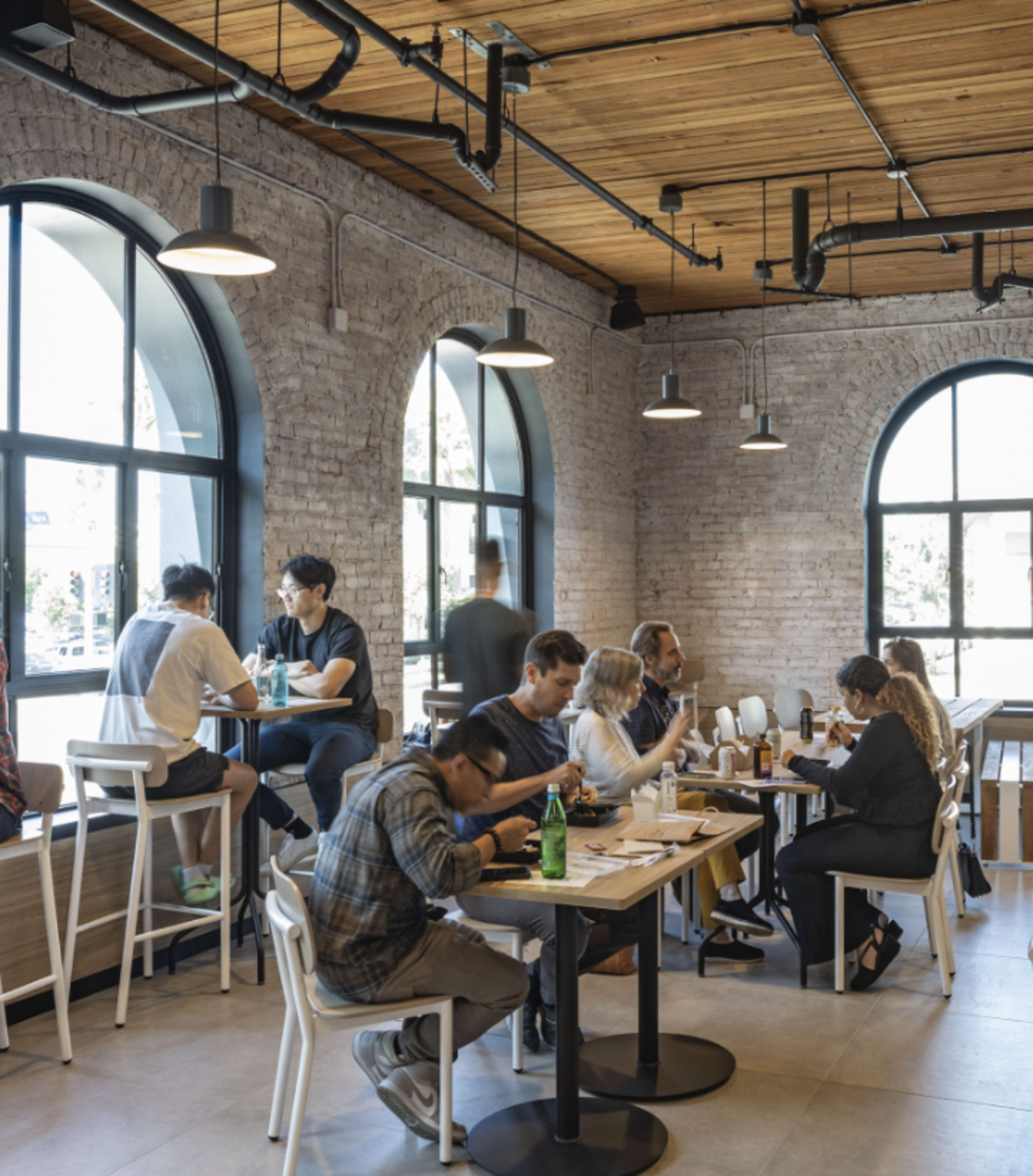 A modern café features groups of people dining and socializing at various tables against a backdrop of brick walls and large arched windows.