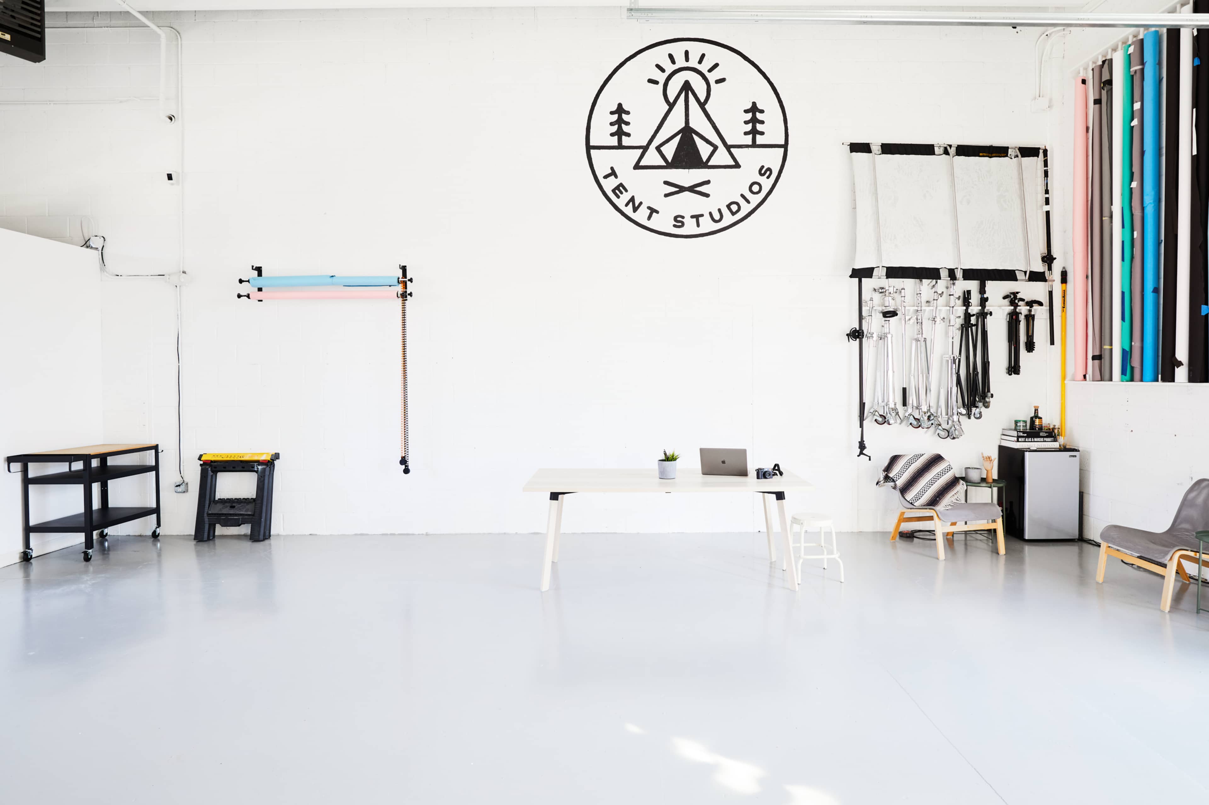 A minimalist studio space with a white wall featuring a logo, a table with a laptop, and various studio equipment along the walls.