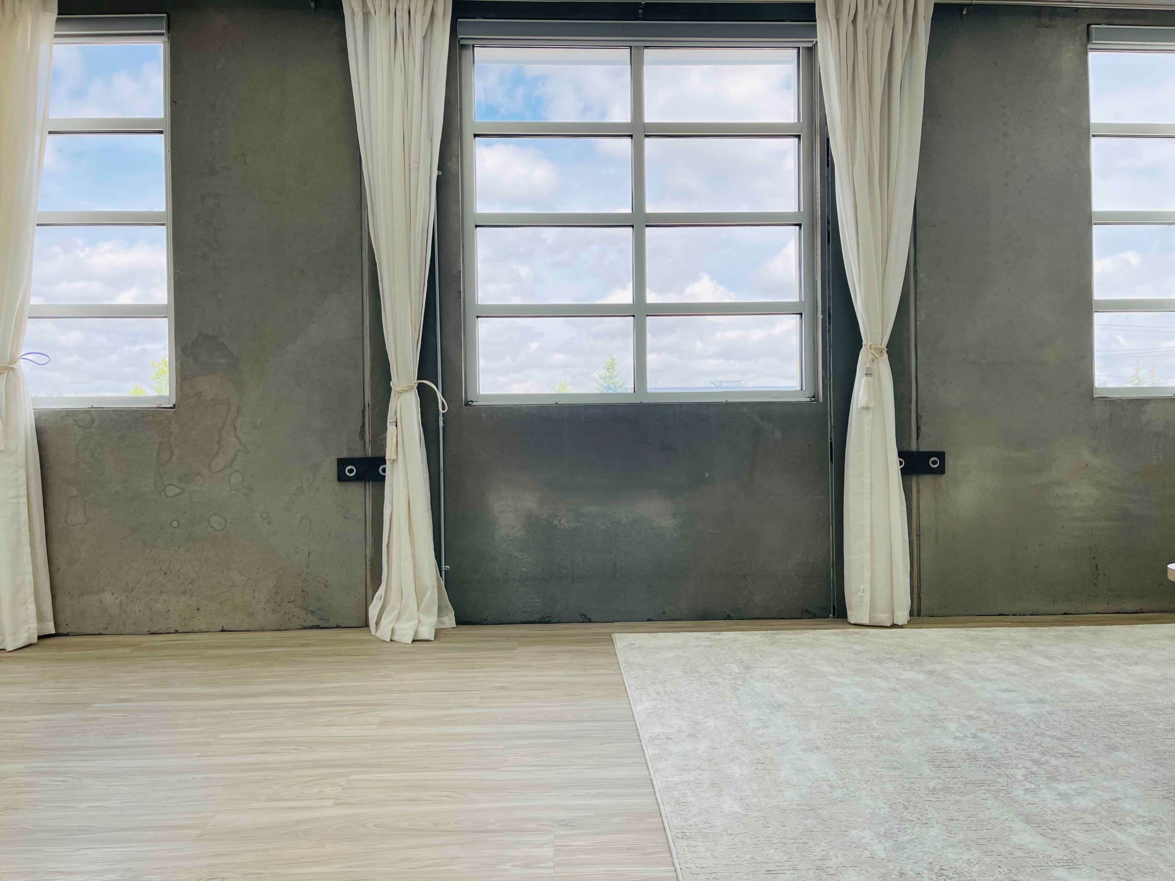 The image shows an interior space with large windows, light-colored curtains, a wooden floor, and a plain area rug.
