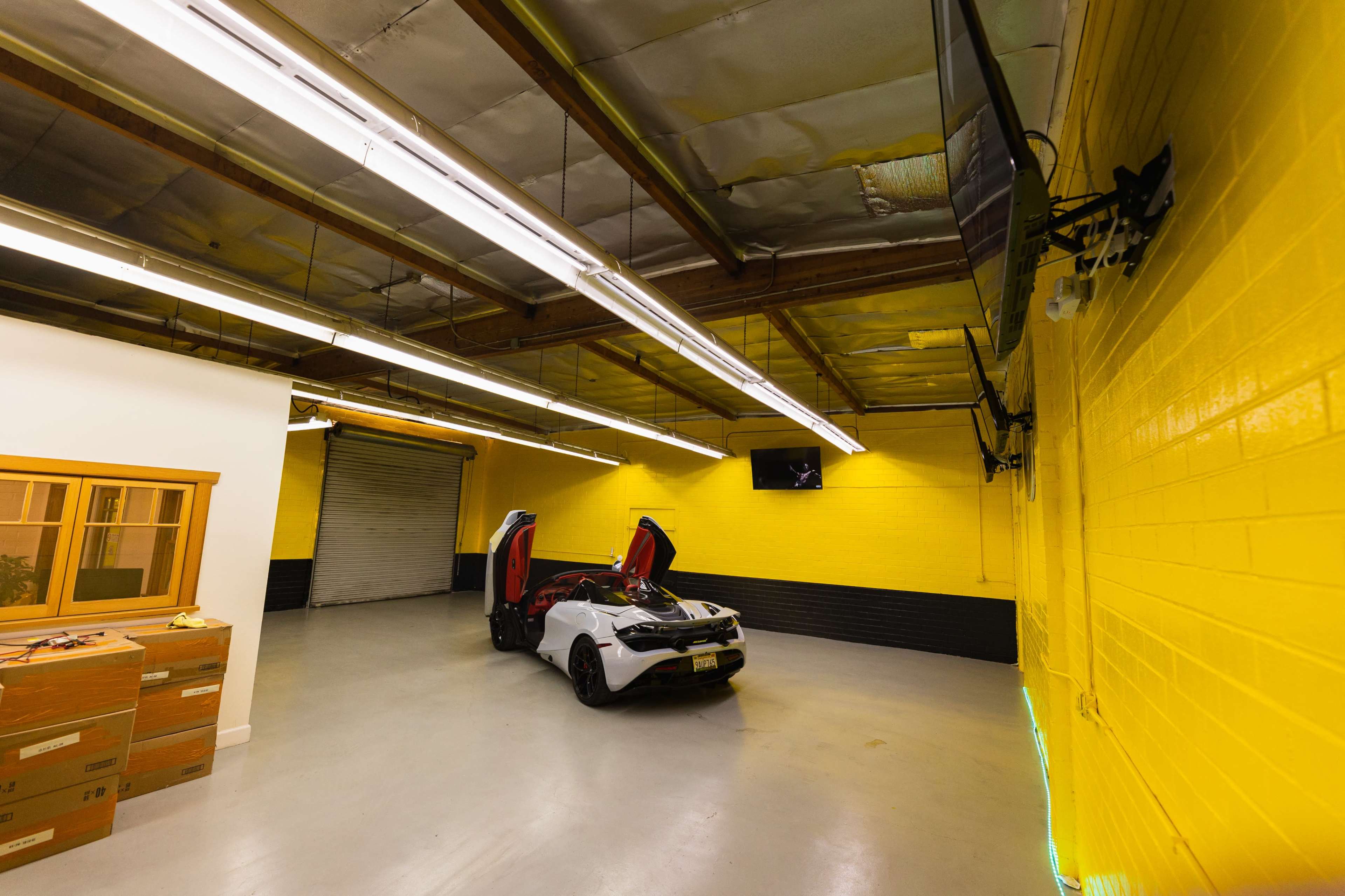 A white sports car with its doors open is parked in a brightly lit garage with yellow walls and a gray floor.