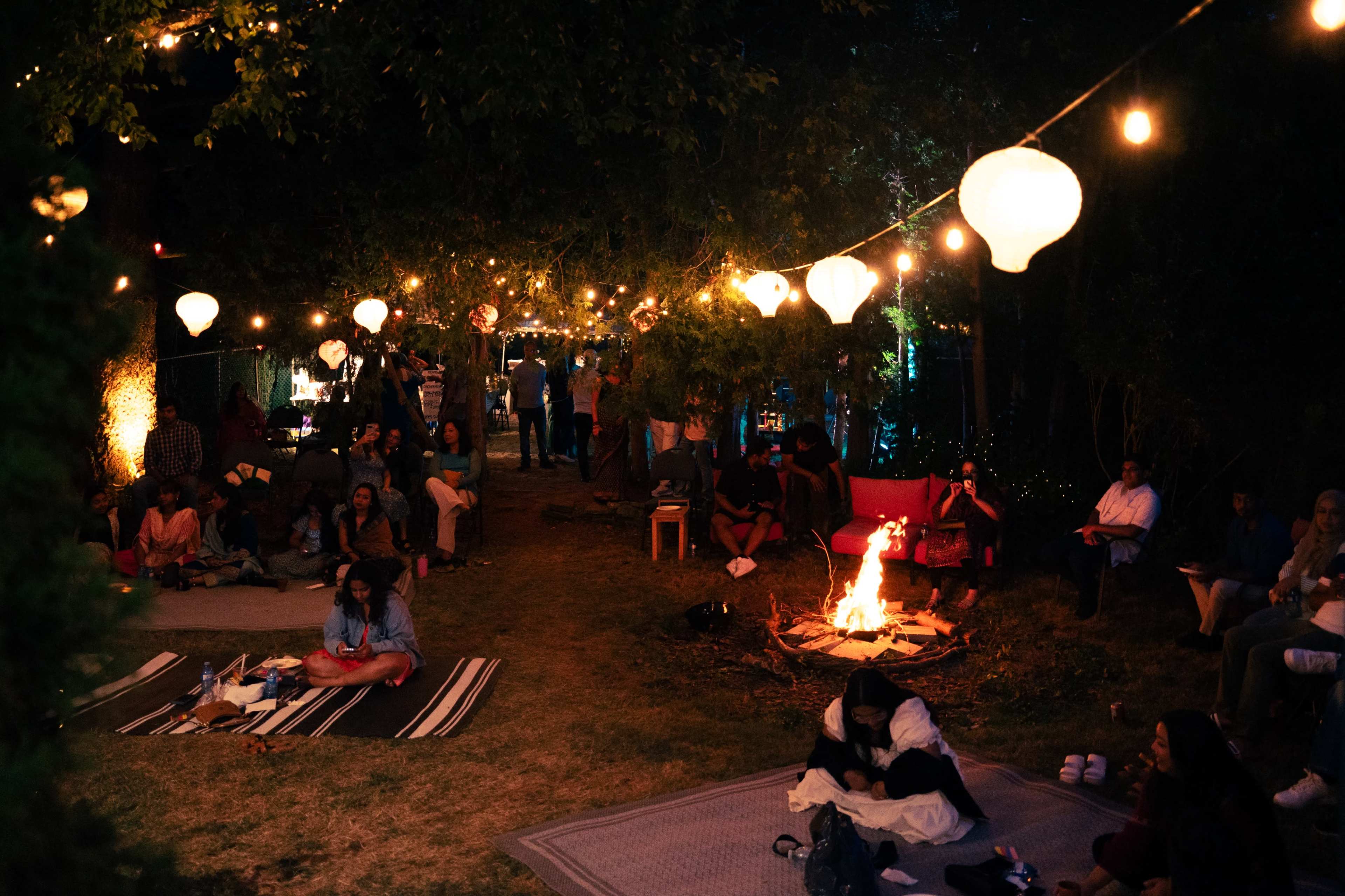 A gathering takes place outdoors at night, with people sitting around a fire and string lights illuminating the area.