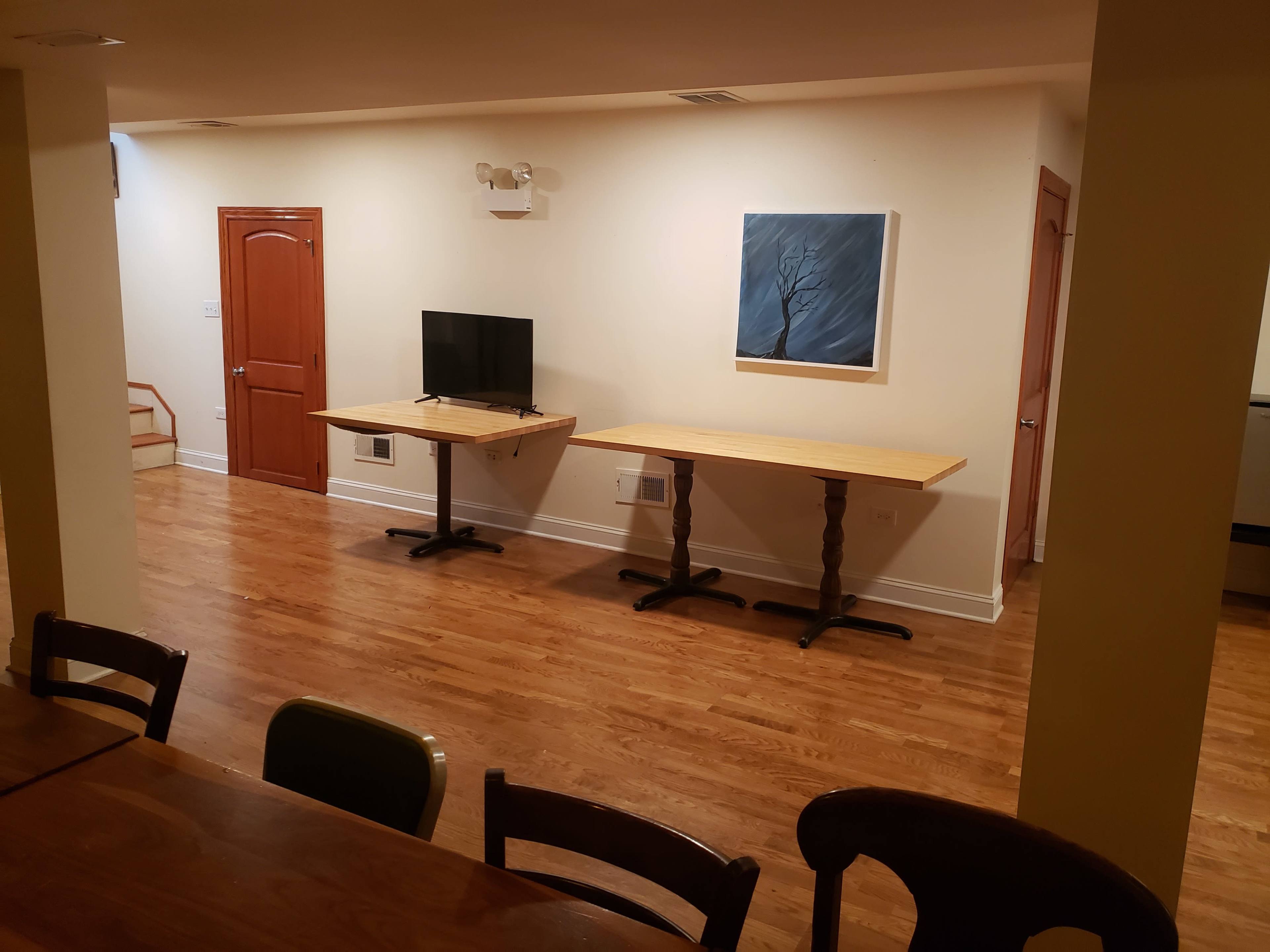 A room with a wooden floor, two tables positioned in the center, a television on one table, and a painting on the wall.