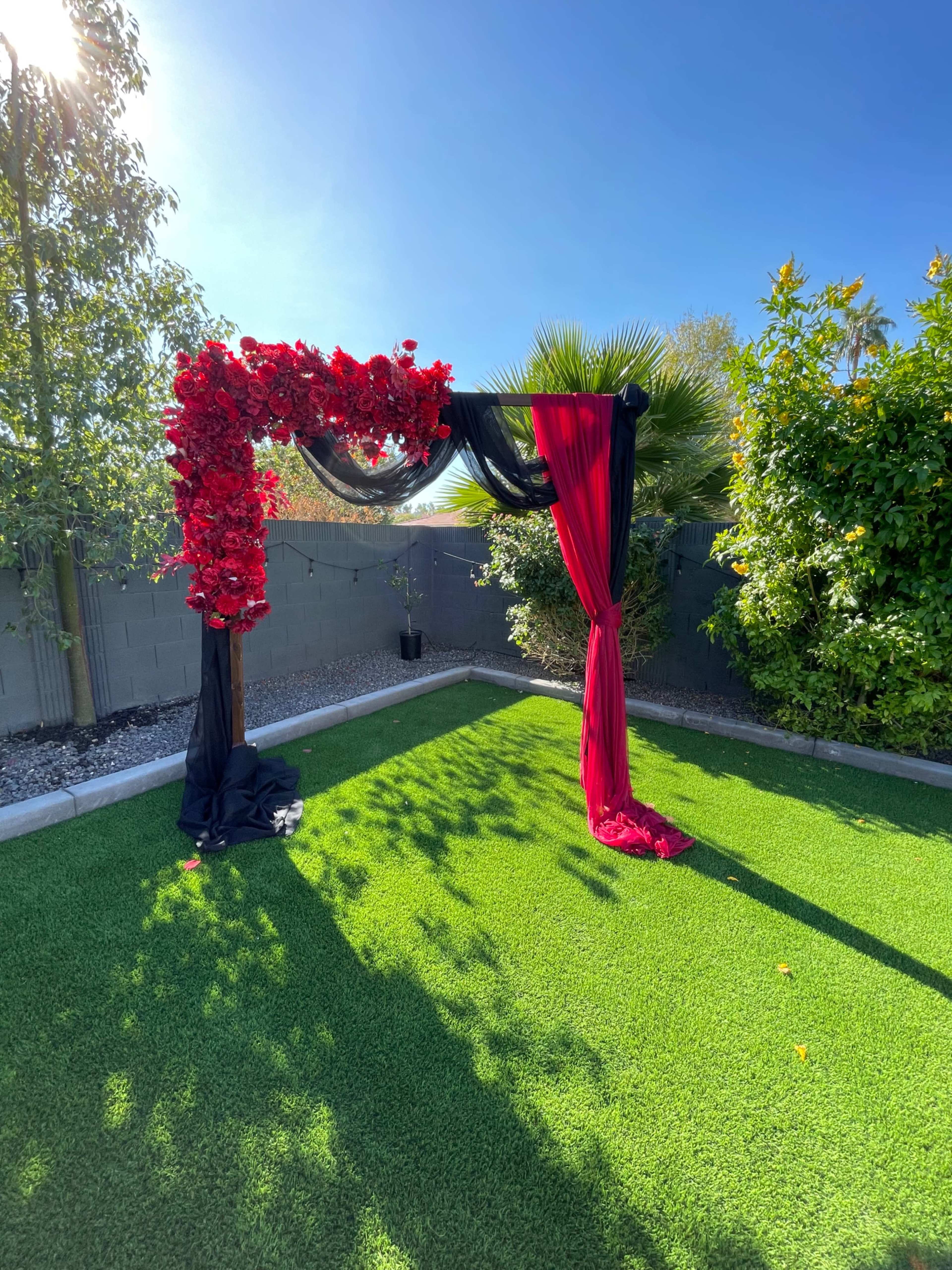 A decorative arch with red flowers and black fabric stands on a grassy area surrounded by greenery under a clear blue sky.