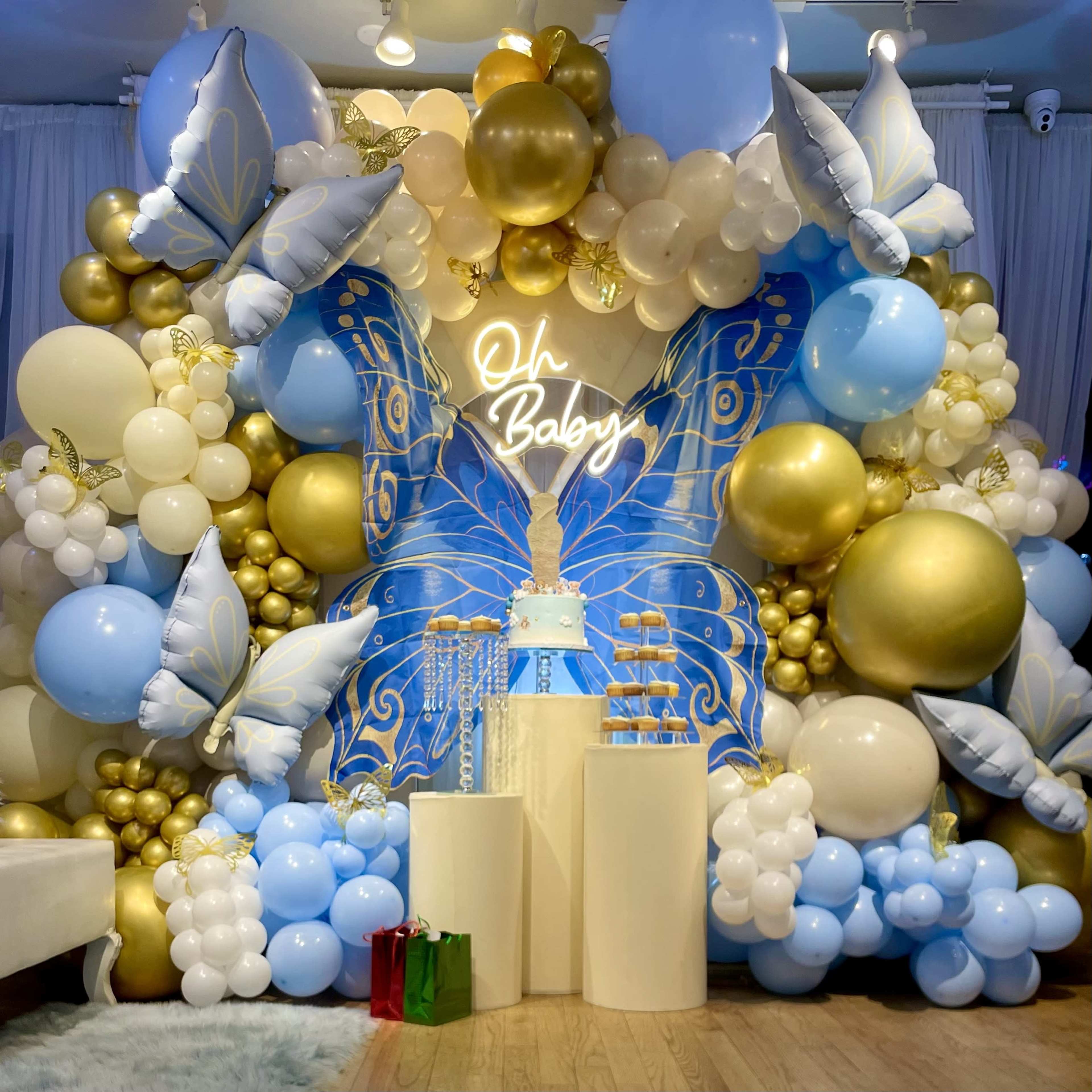 The image shows a festive balloon installation featuring a large butterfly design, a white cake on display, and decorative elements in gold and blue.