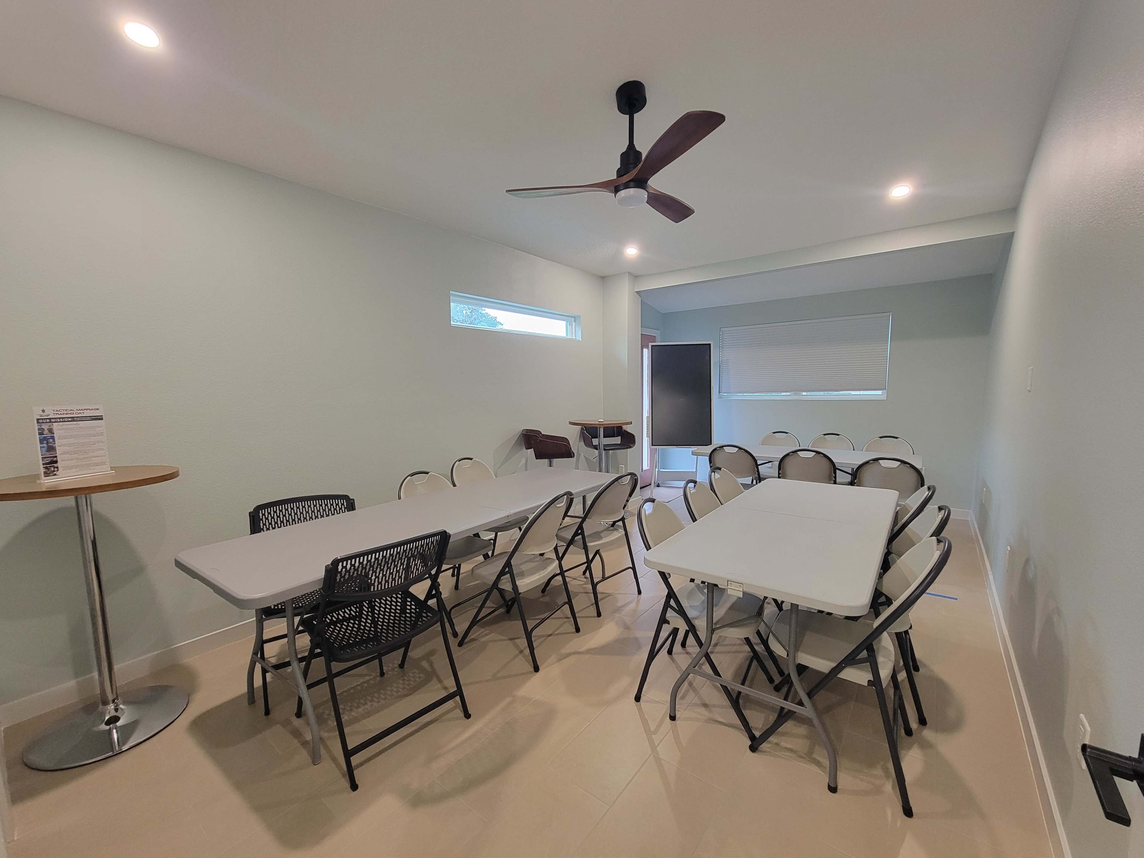 The image shows a light green meeting room with multiple folding tables and chairs arranged neatly, along with a ceiling fan and a whiteboard in the background.