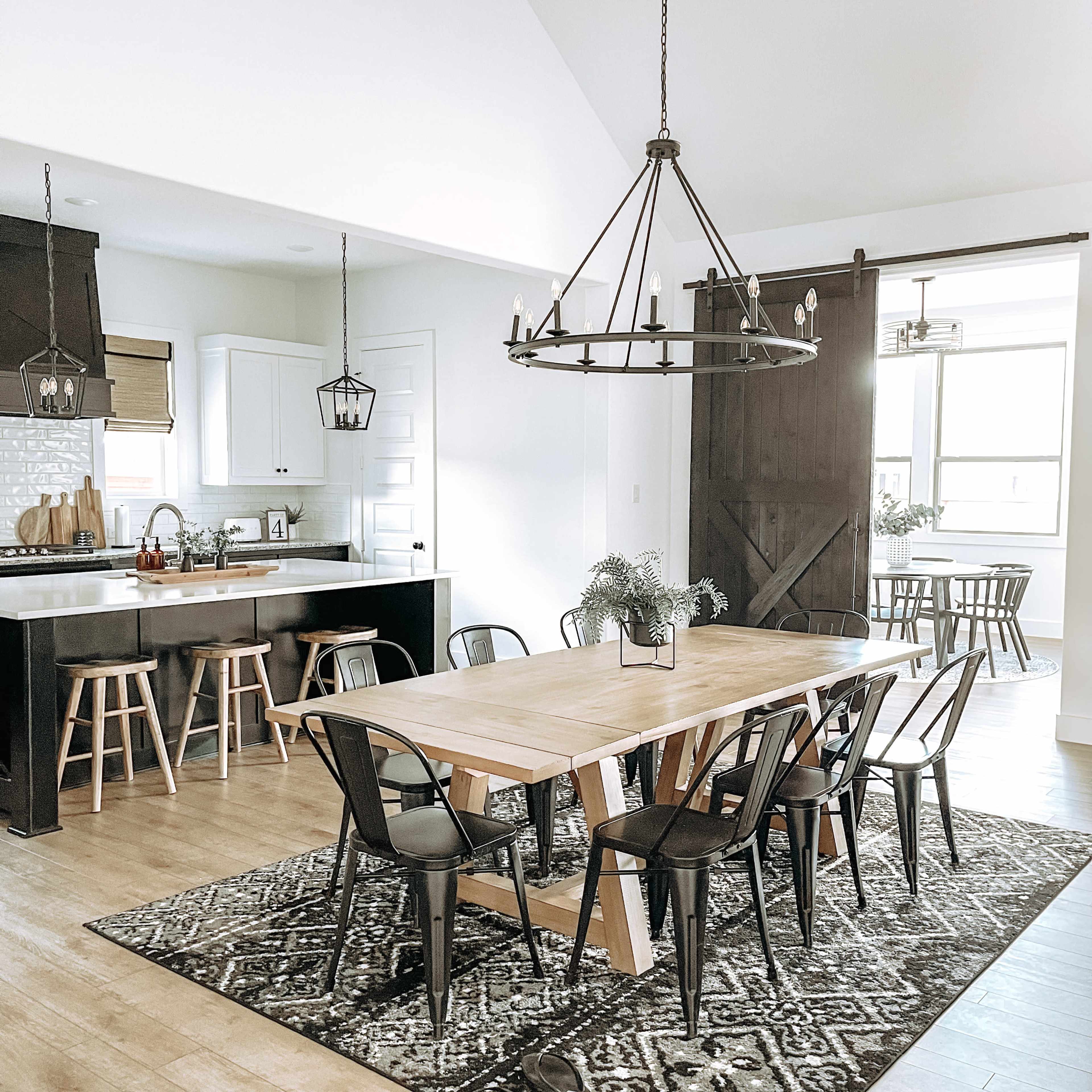 A modern dining area with a wooden table surrounded by black chairs, a light fixture overhead, and a view of a kitchen area in the background.
