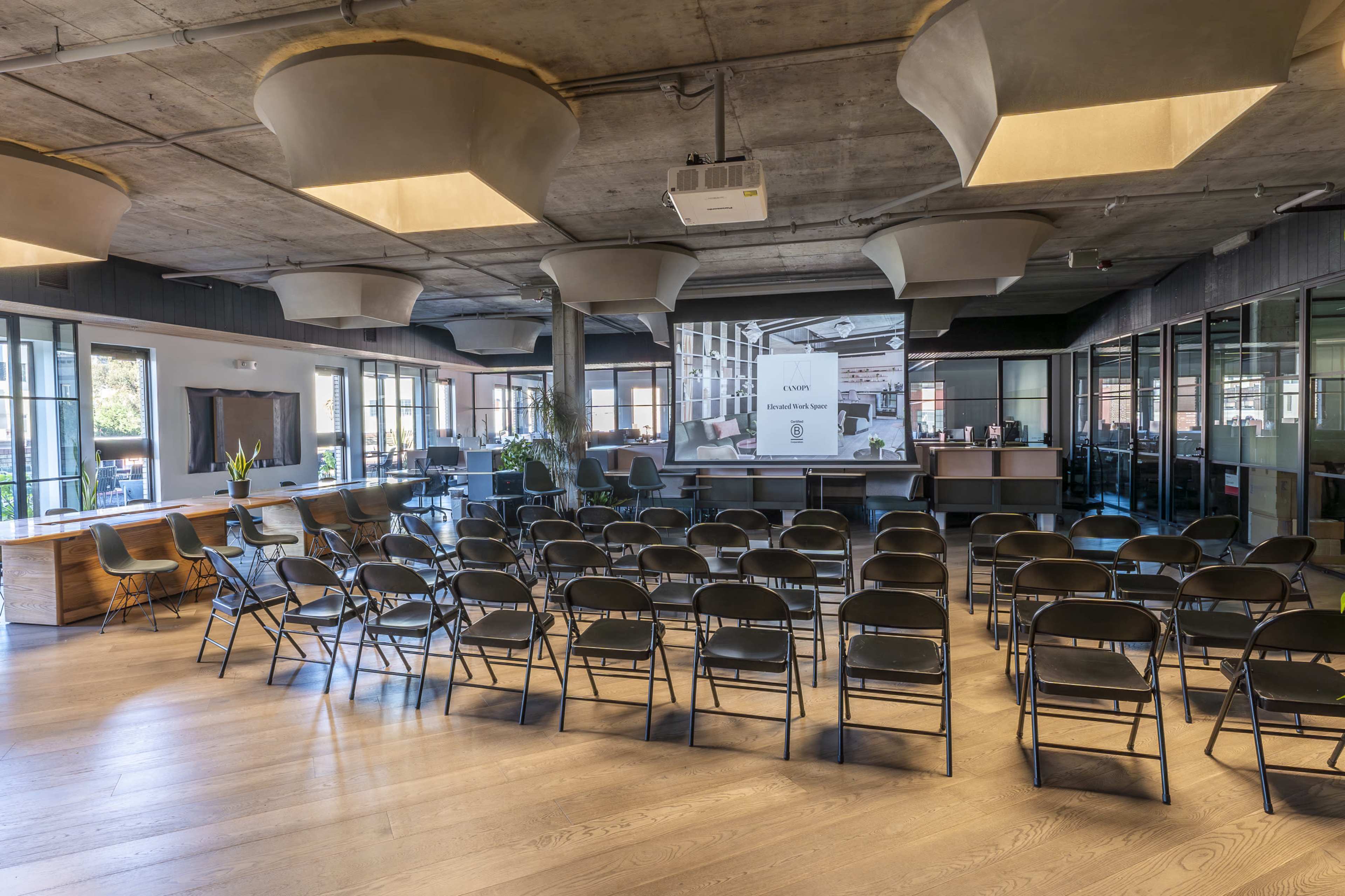 The image shows a modern event space with rows of black chairs facing a projector screen, surrounded by large windows and a minimalist design.