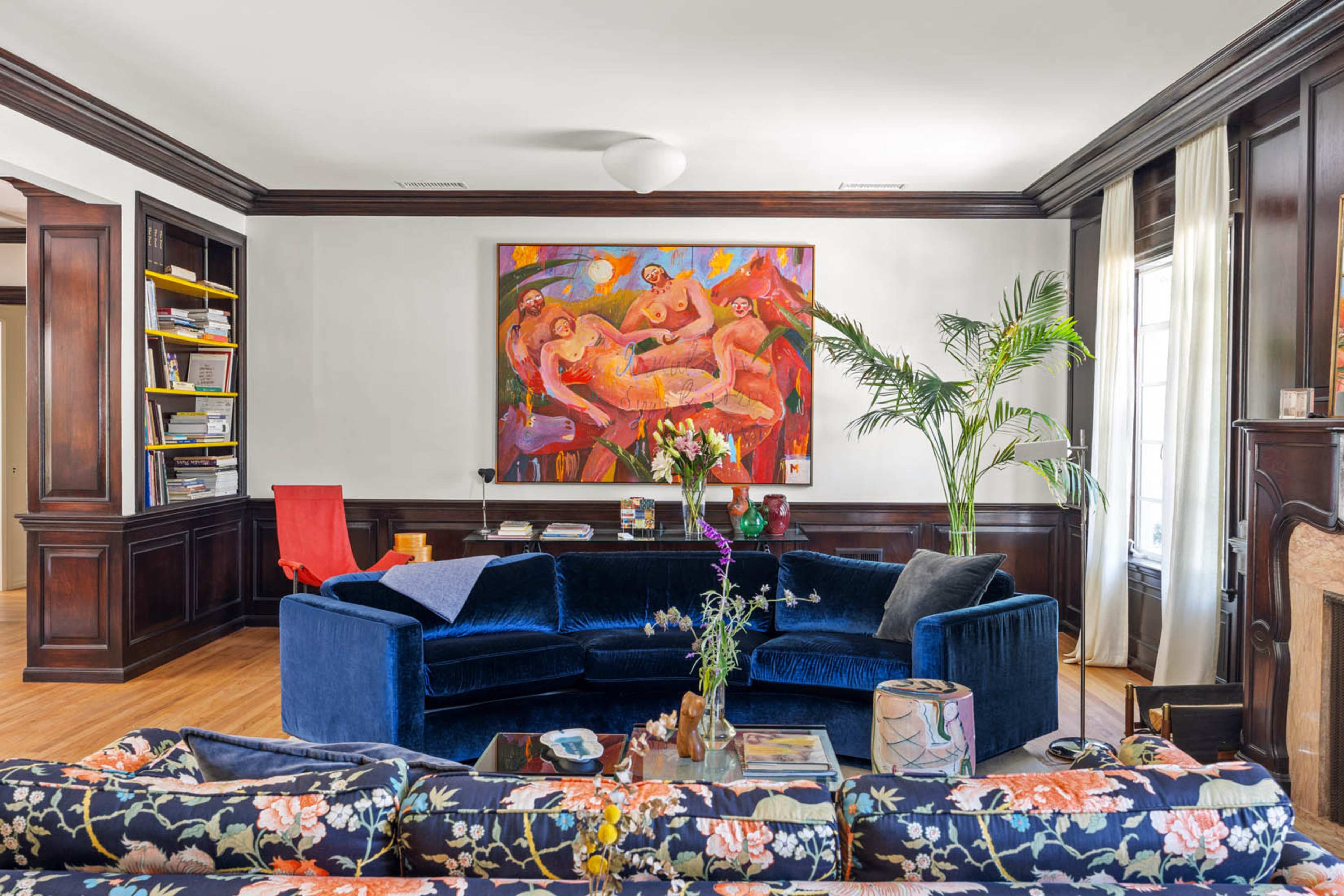 The living room features a large blue sectional sofa centered around a painted wall adorned with a colorful artwork, flanked by wooden bookshelves and a potted plant.