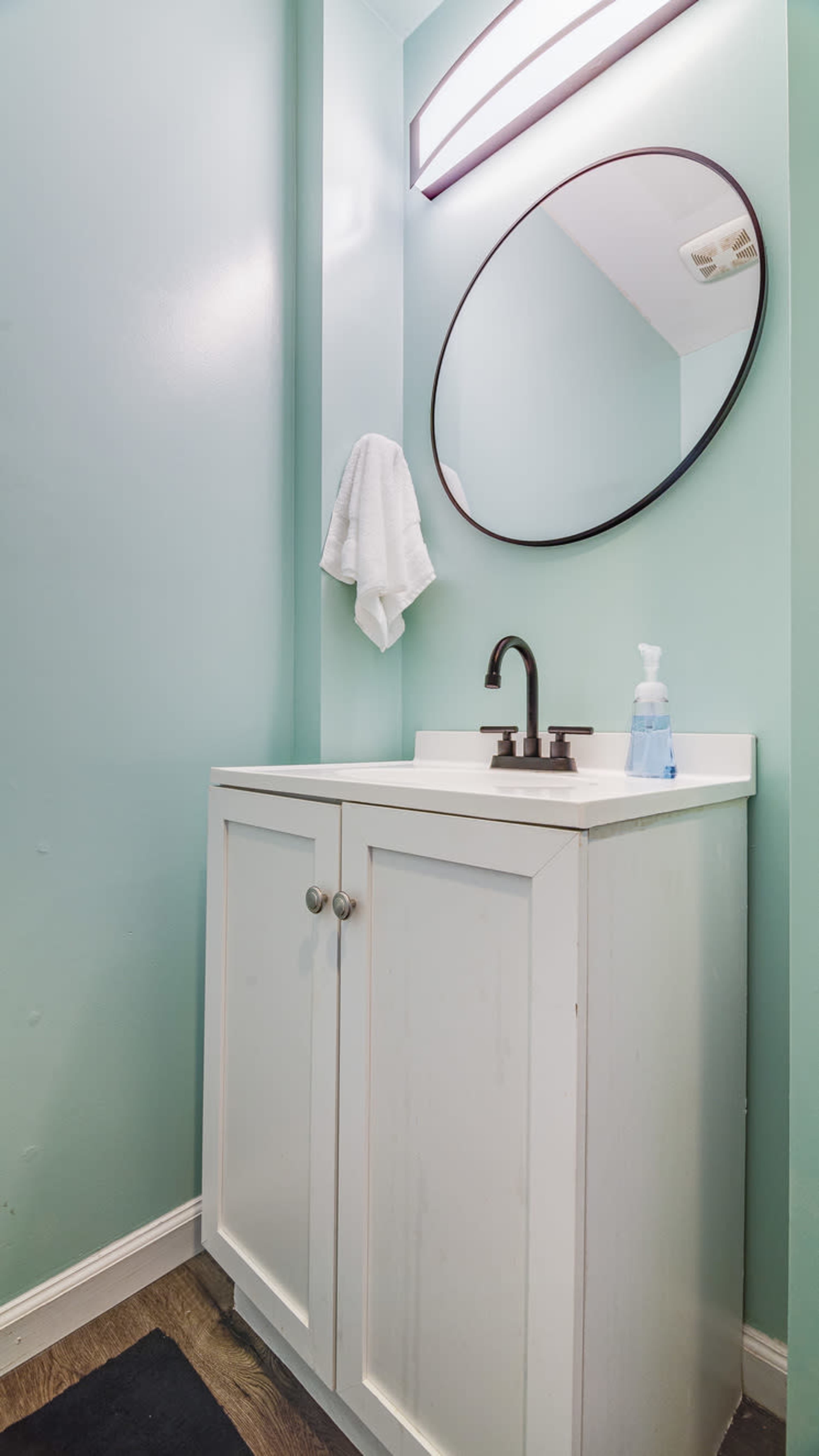 A small bathroom features a light blue wall, a white cabinet with a sink, a round mirror above it, and a towel hanging nearby.