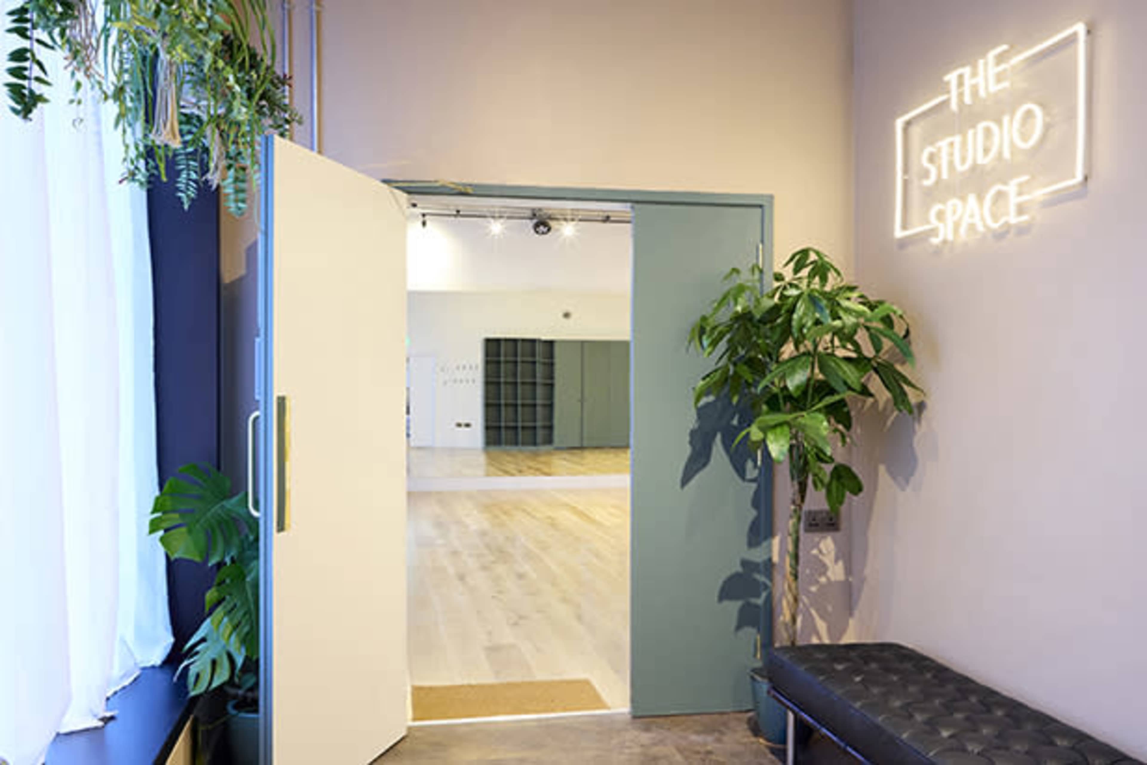 A brightly lit entryway leads into a spacious studio area, featuring large mirrors and wooden flooring, with a neon sign reading "THE STUDIO SPACE" on the wall.
