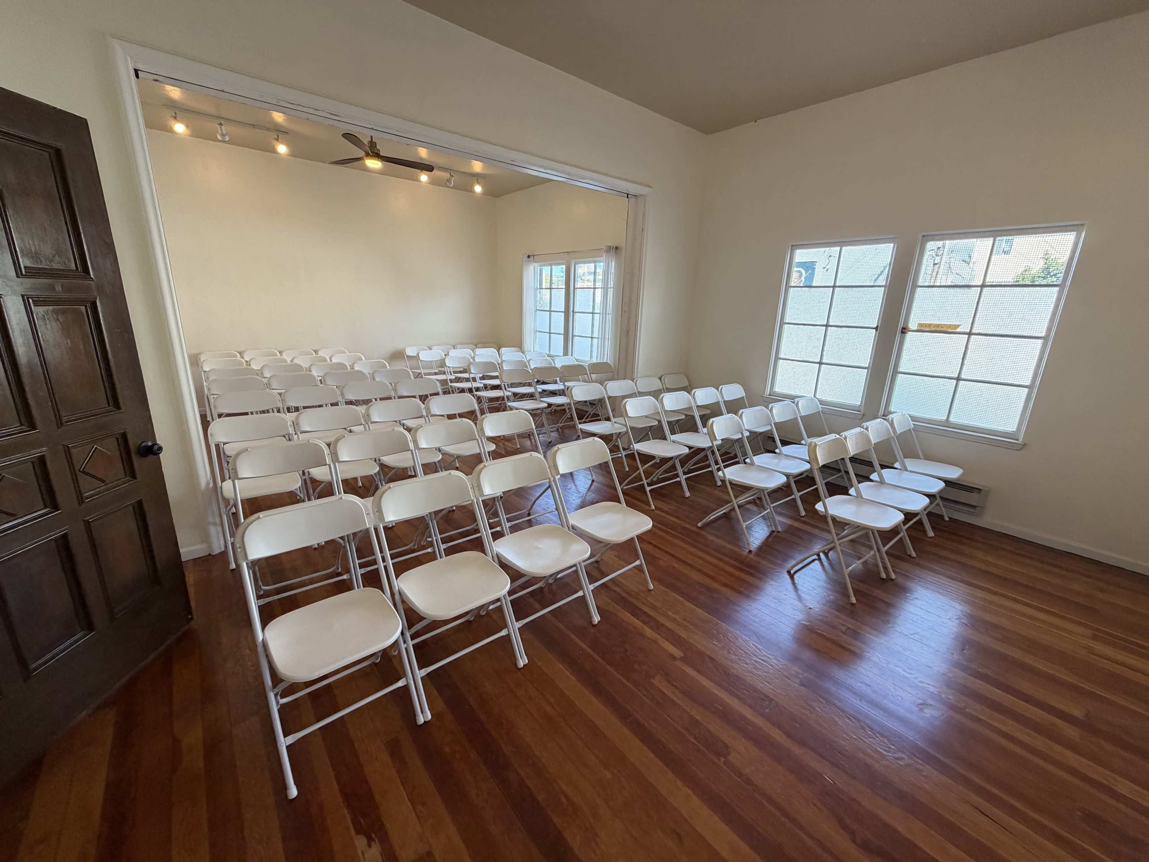 The image shows a room arranged with several rows of white chairs on a wooden floor, facing a brightly lit window.