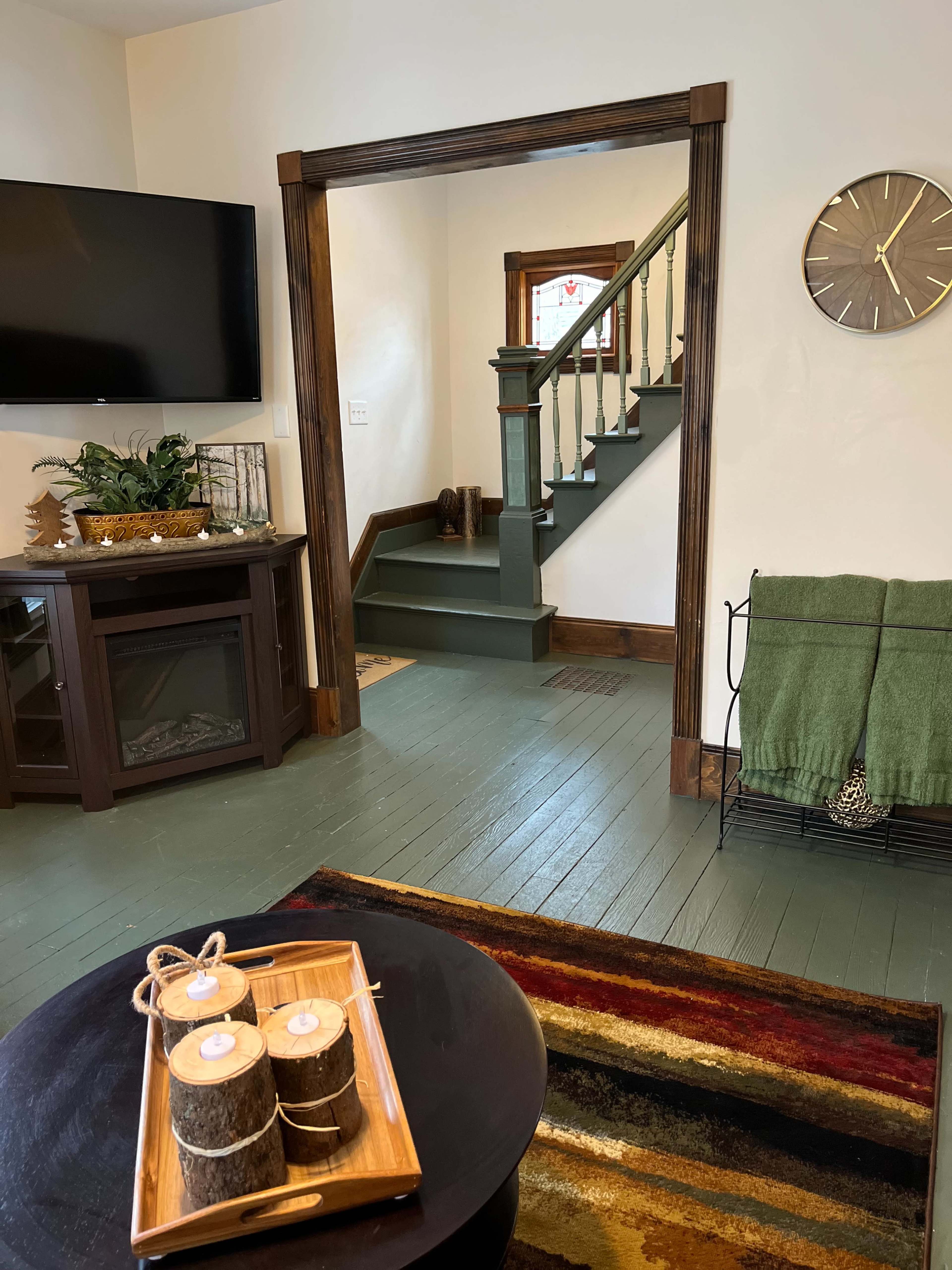 A living room with a black coffee table displaying candles, a TV mounted on the wall, a staircase leading to an upper level, and a green throw blanket draped over a metal rack.