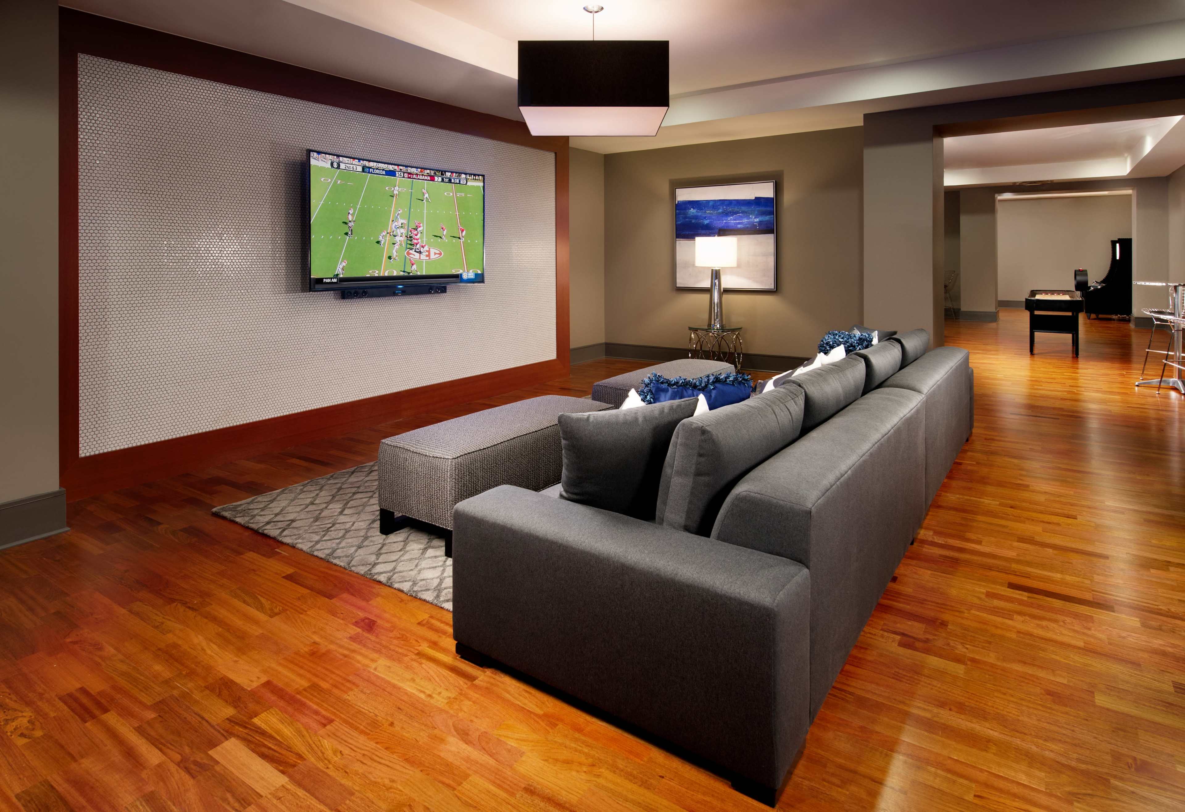 A modern living room features a gray sectional sofa facing a wall-mounted television showing a sports game, with wooden flooring and soft lighting.
