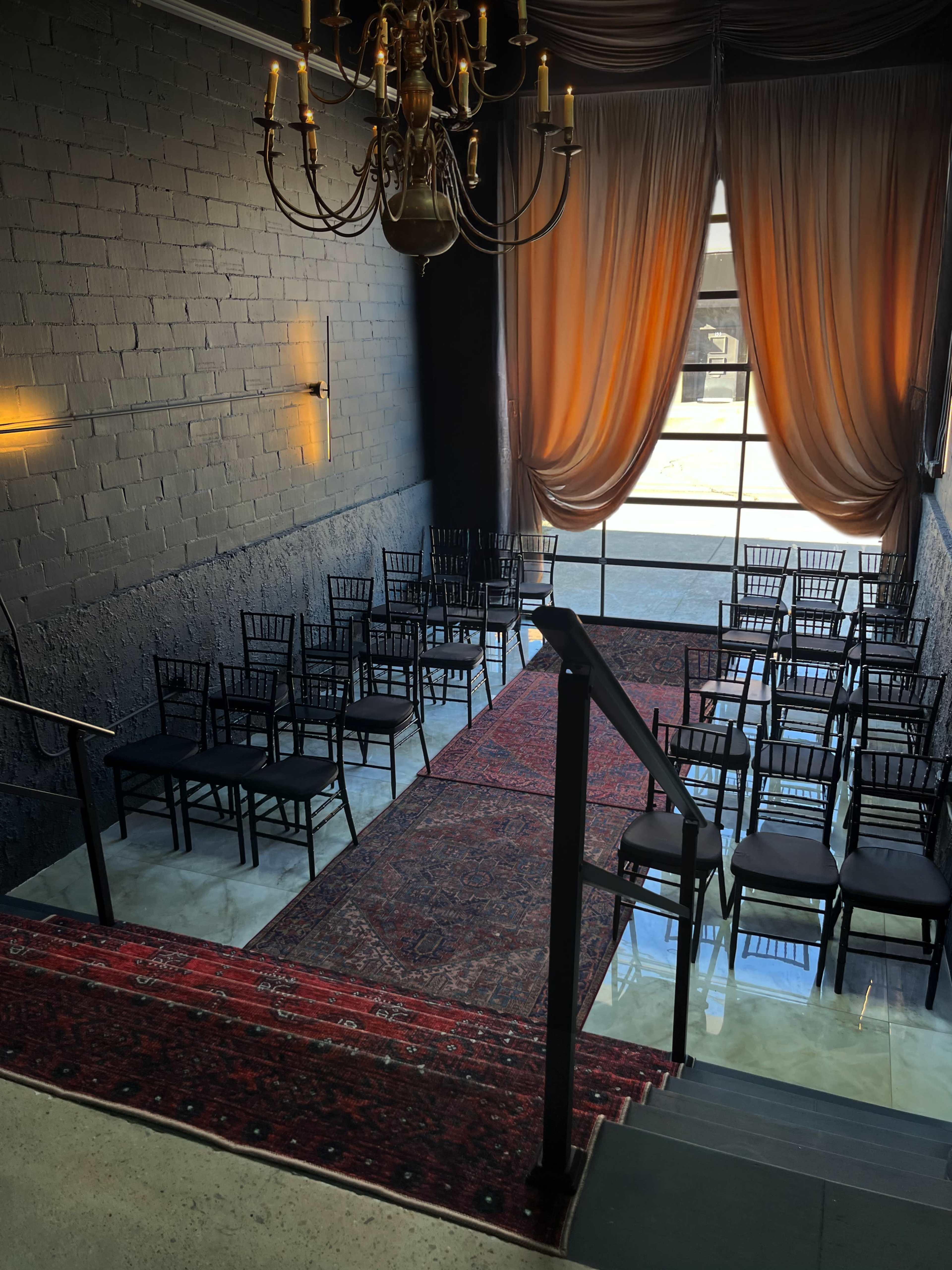 The image shows an indoor venue featuring a chandelier, a large window, and rows of black chairs arranged in front of a red rug.