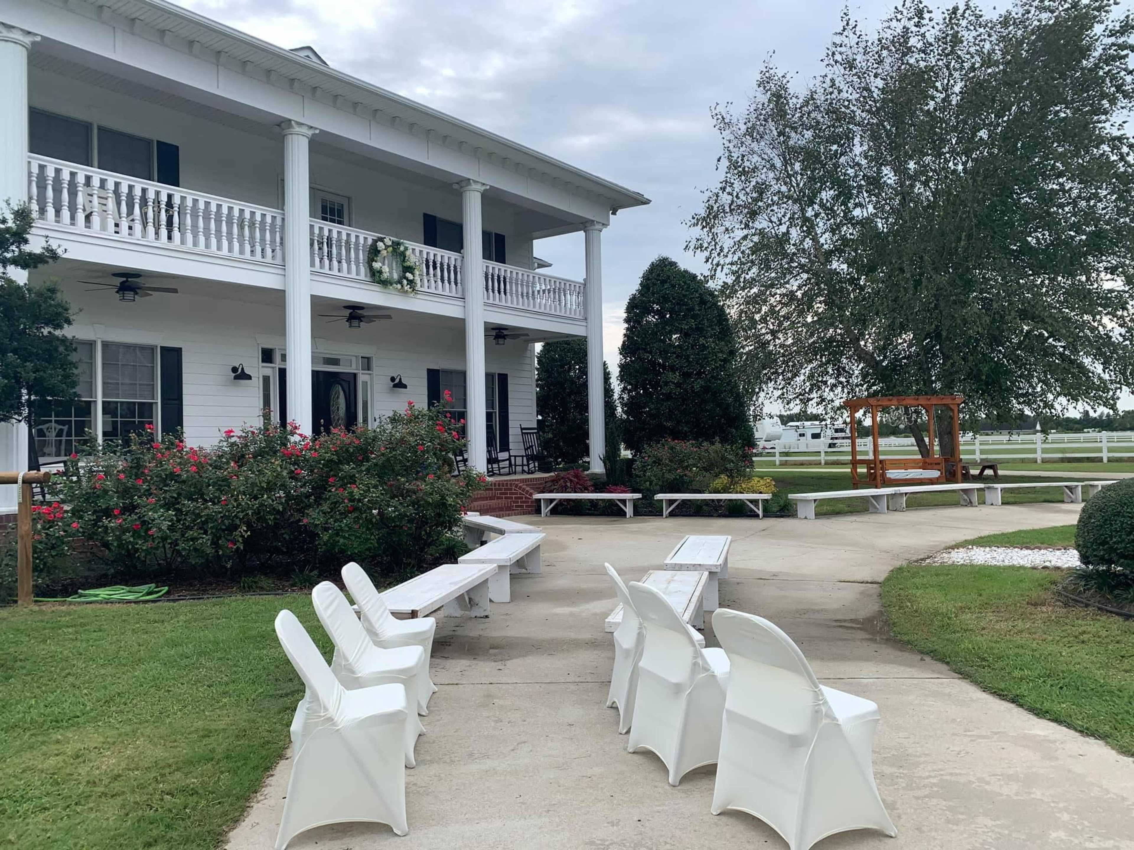 A white house with a large porch and multiple columns is surrounded by green grass, benches, and a swing set in the yard.