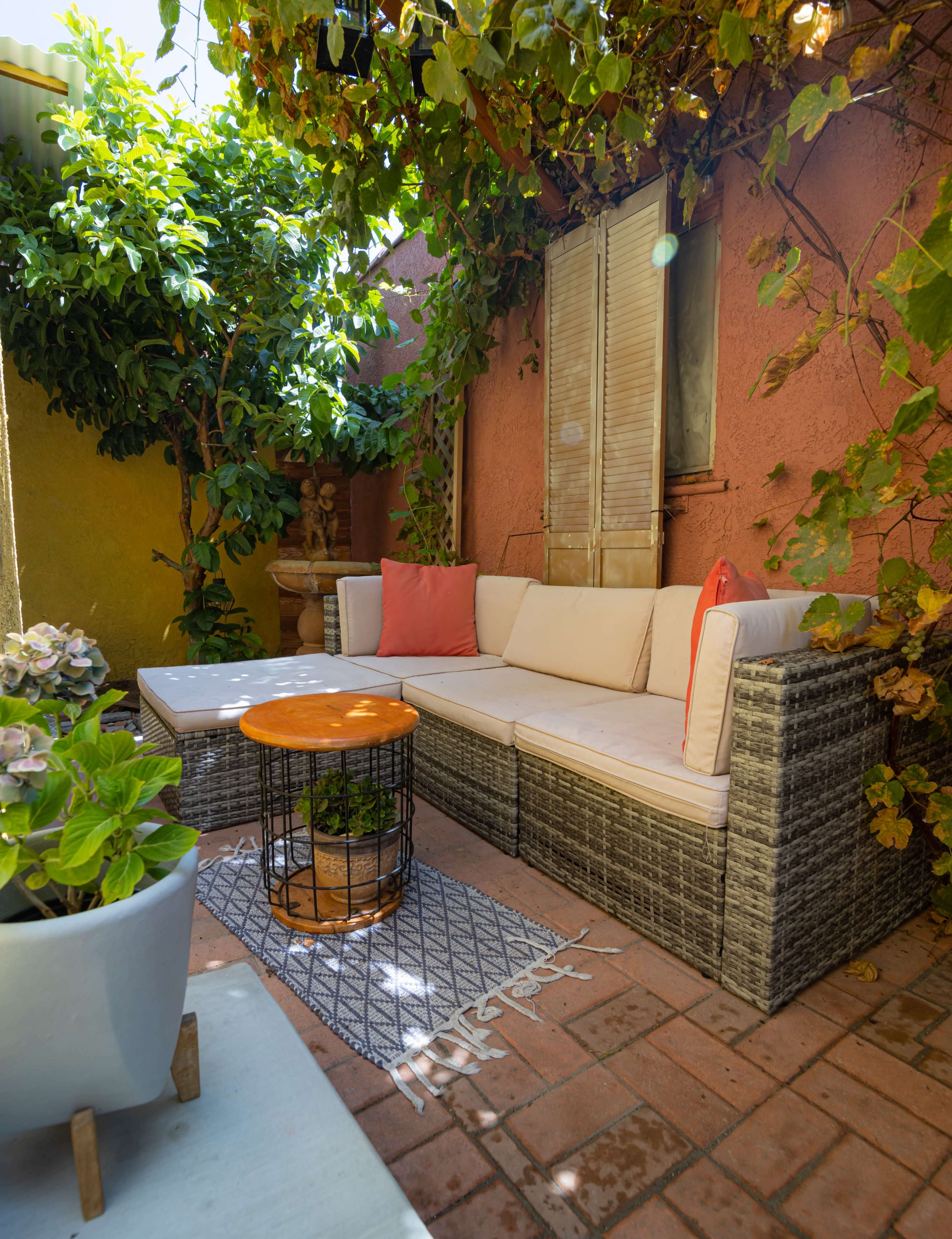 A cozy outdoor seating area features a wicker sofa with cushions, a wooden coffee table, and surrounding greenery against a colorful wall.