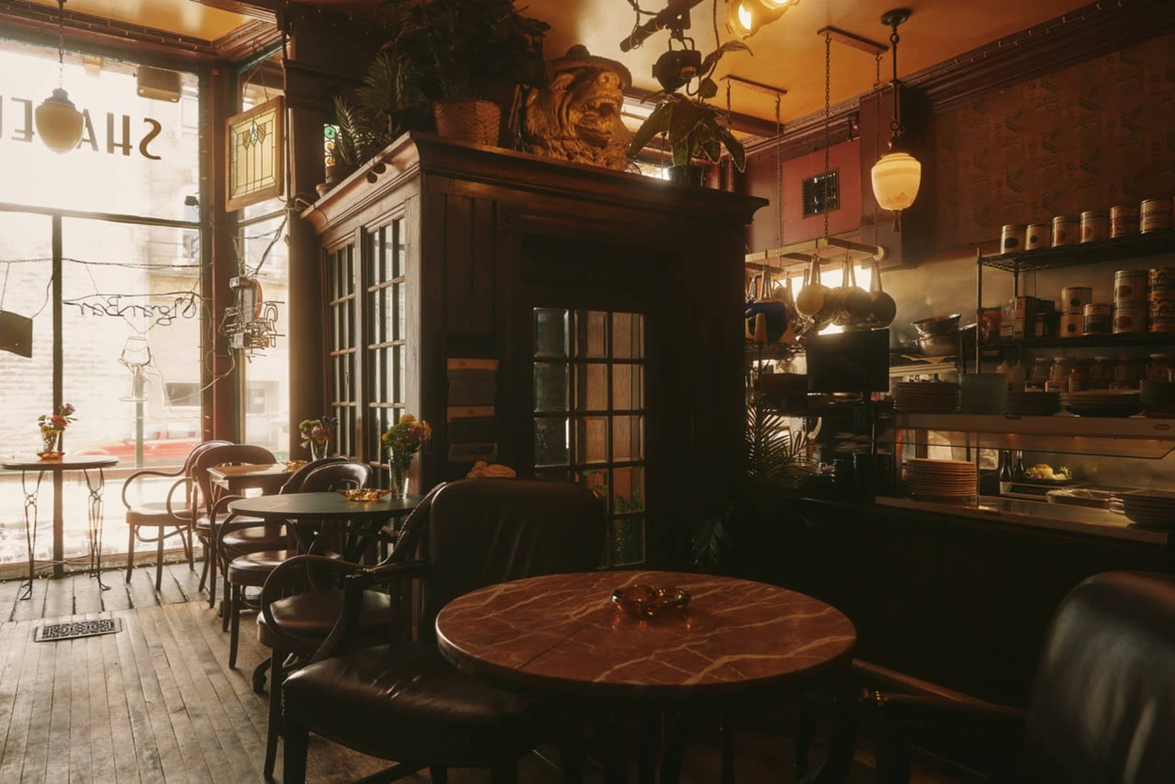 The image shows the interior of a cozy café with wooden furniture, a large window, and various plants decorating the space.