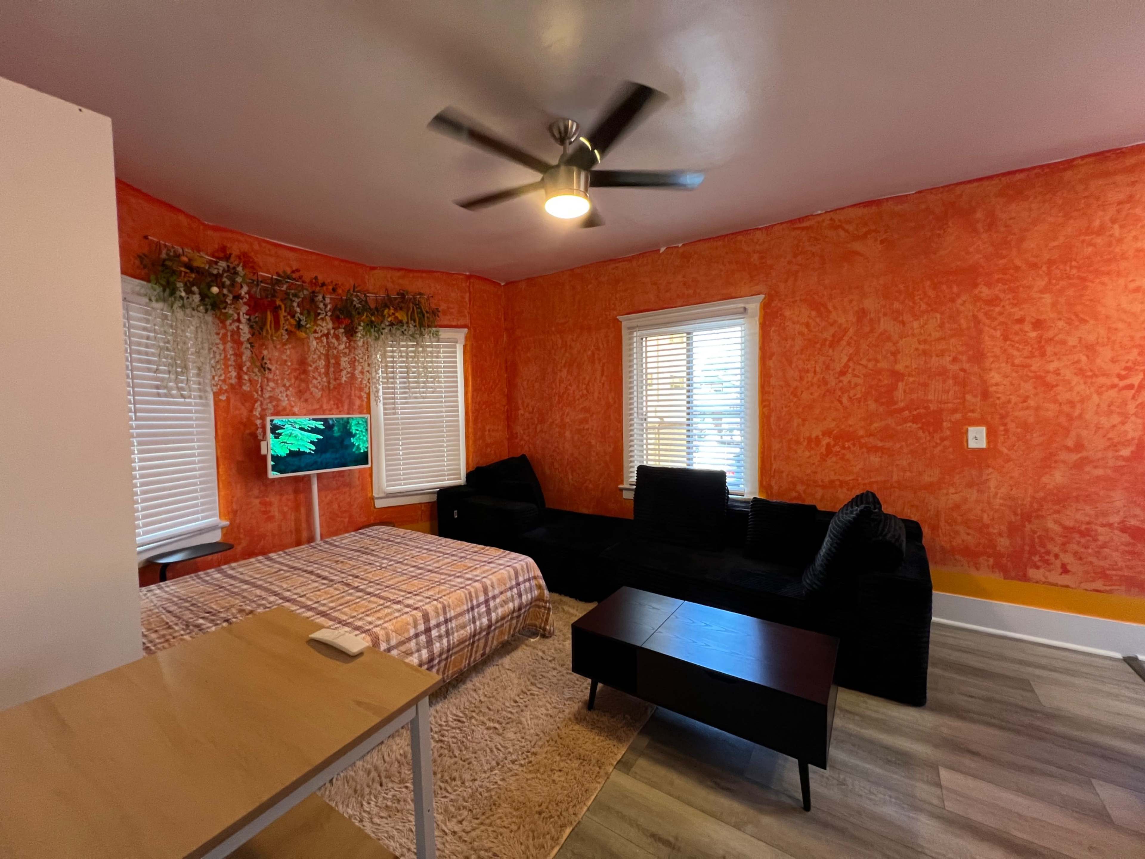 The image shows a room with an orange textured wall, a bed with a plaid blanket, a black sofa, and a small table beside a television on the wall.