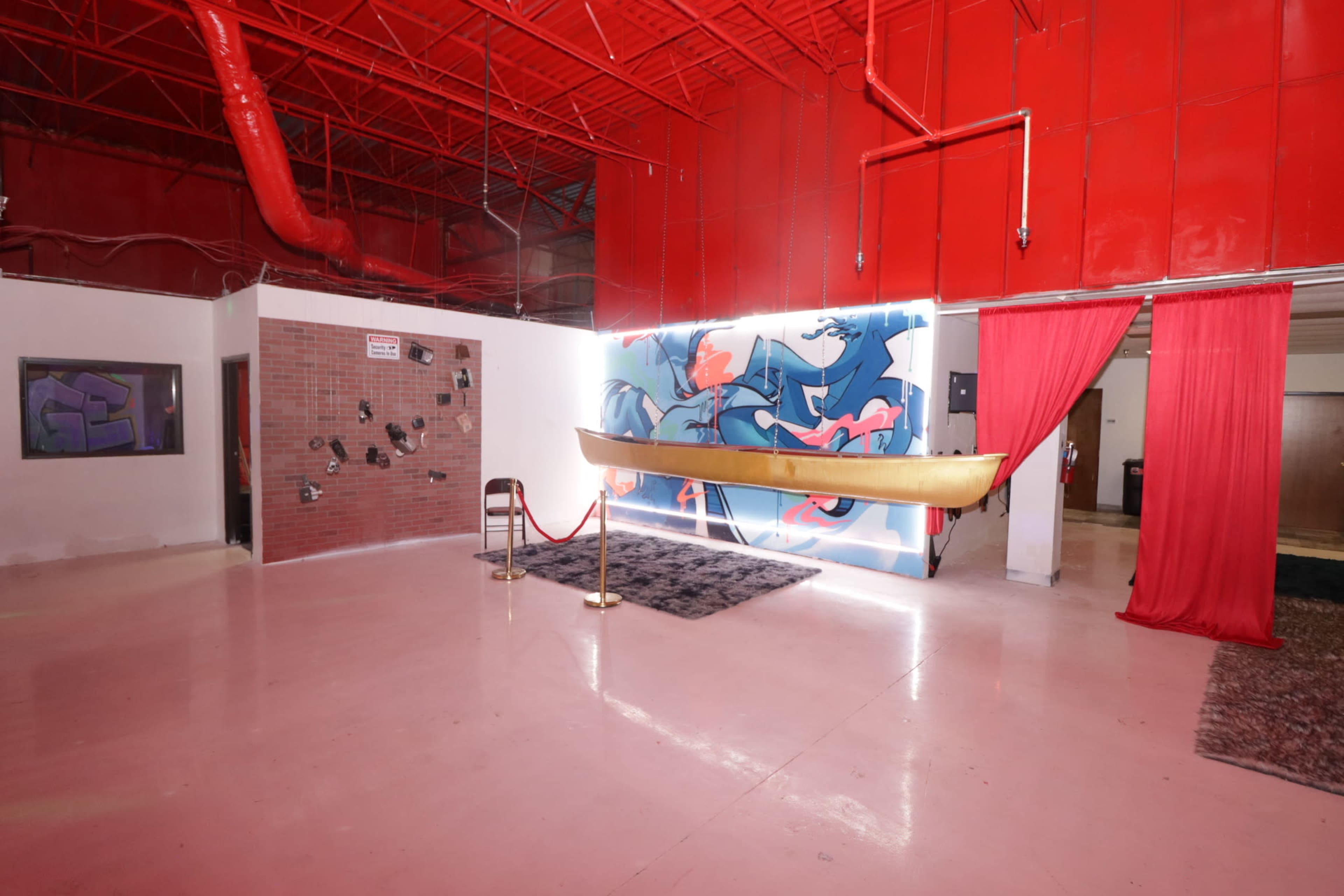 The image depicts an interior space with a bright red ceiling, a mural of blue figures on the wall, a suspended canoe, and red curtains framing the area.
