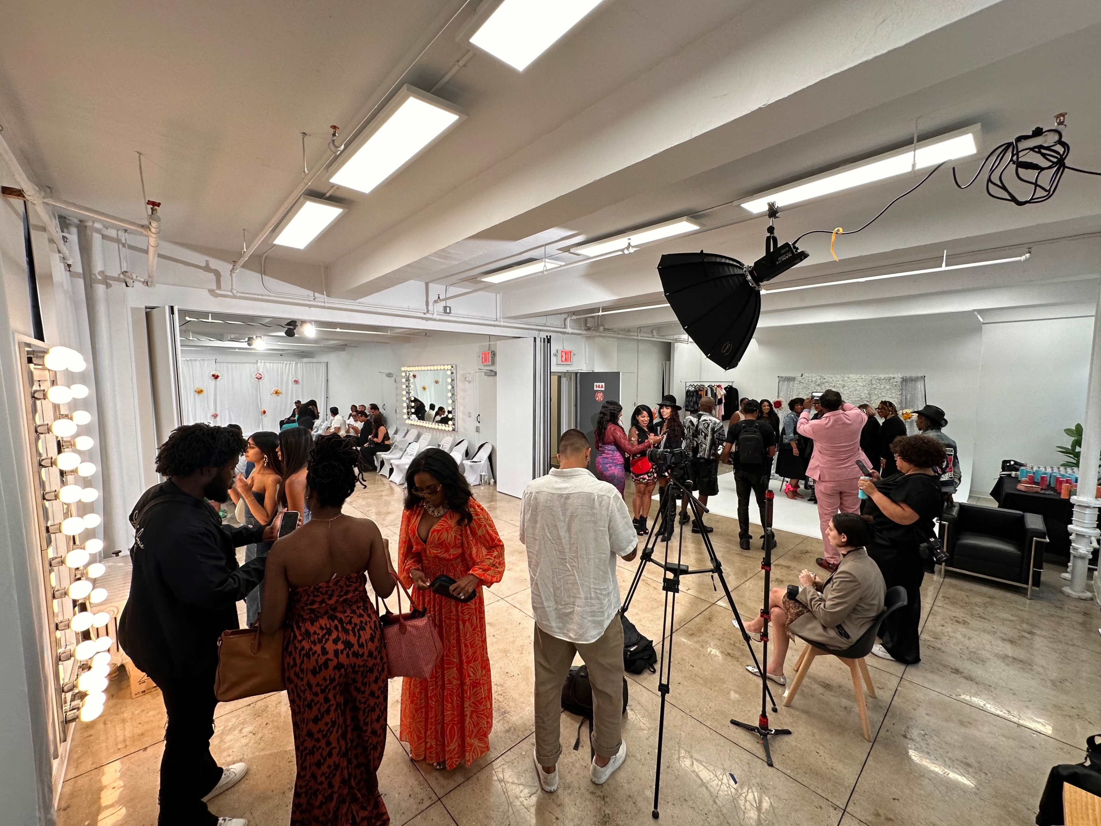 A crowded studio space features people socializing, taking photos, and preparing for a creative event under bright overhead lights.