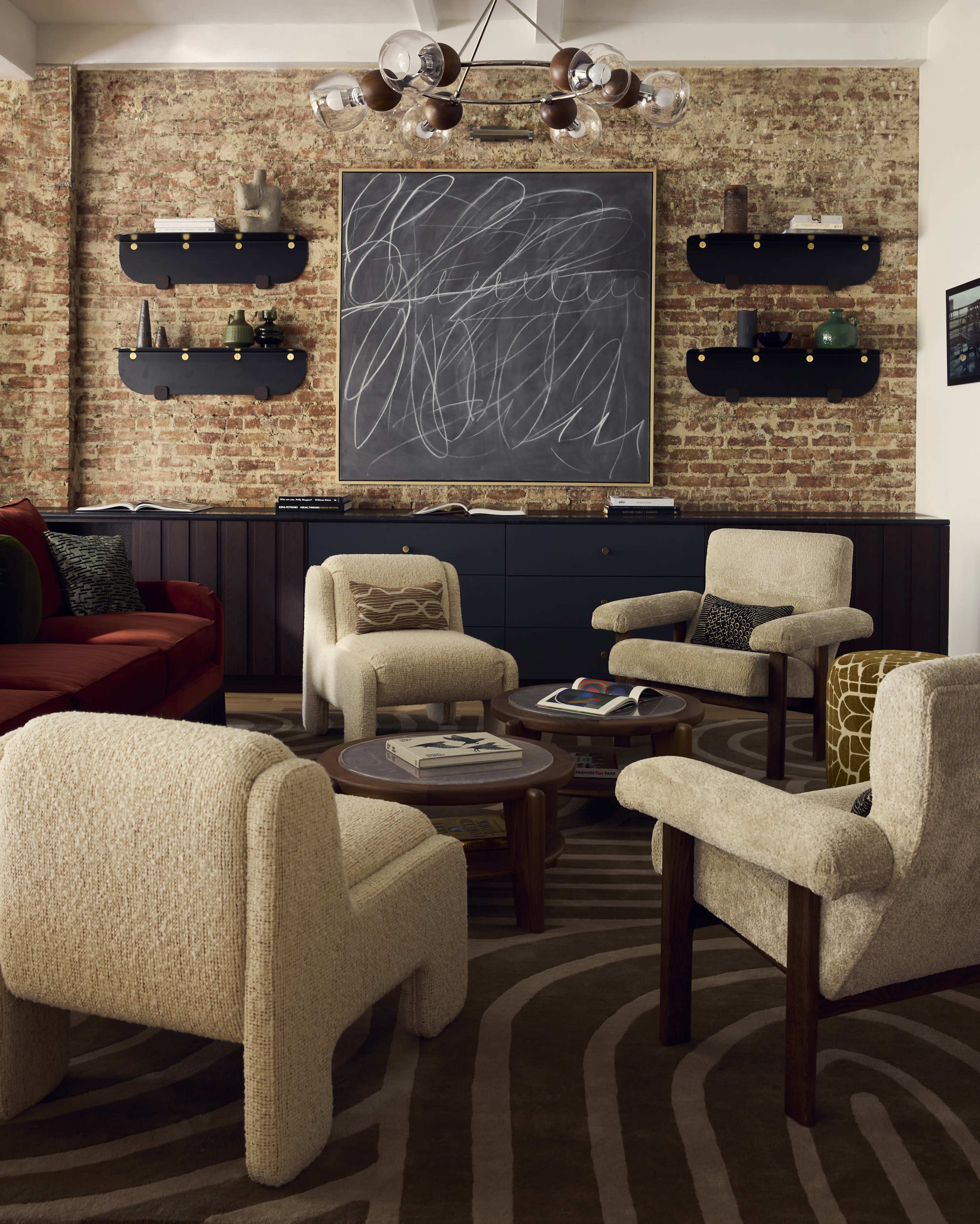 The image shows a modern living room with a brick wall, four upholstered chairs around a coffee table, and a large chalkboard featuring abstract drawings above a low cabinet with decorative items.