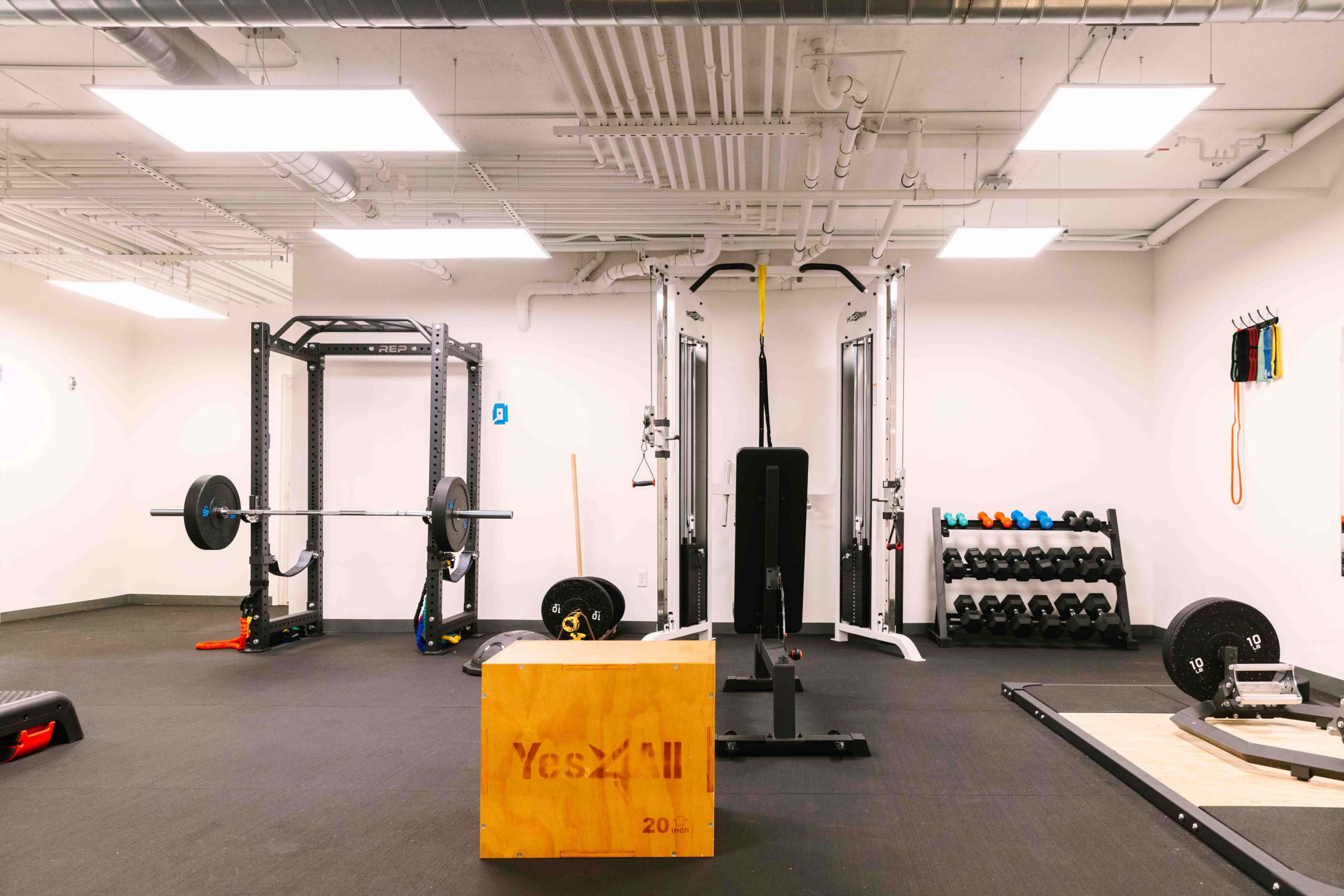 An organized gym space with various weightlifting equipment, including a squat rack, dumbbells, and a wooden box for jump exercises.