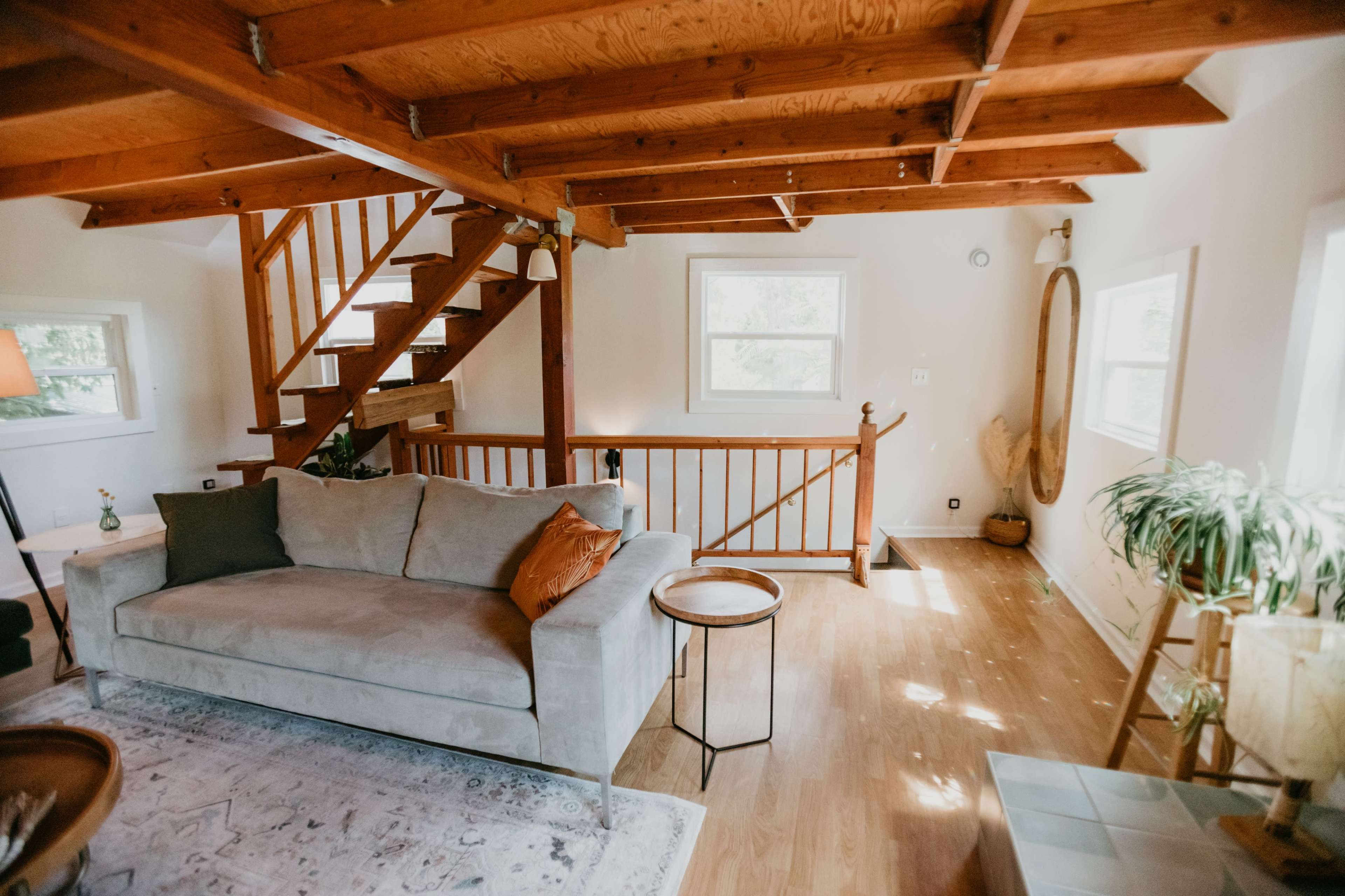 A living room with a light gray sofa, a small round table, wooden beams in the ceiling, and a staircase leading to an upper level.