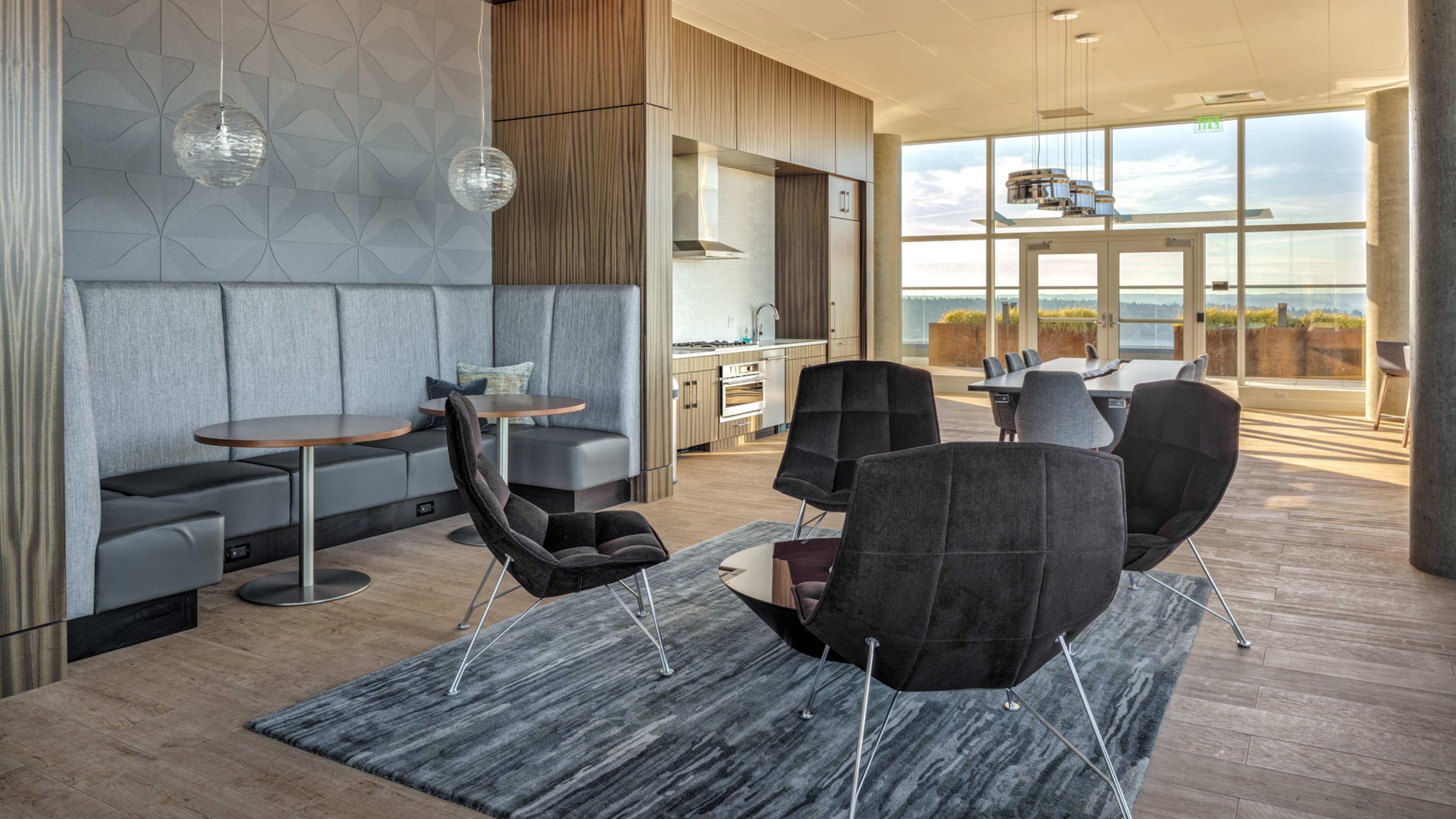 A modern lounge area featuring a dining table, several chairs, a kitchen in the background, and large windows that allow natural light to enter.