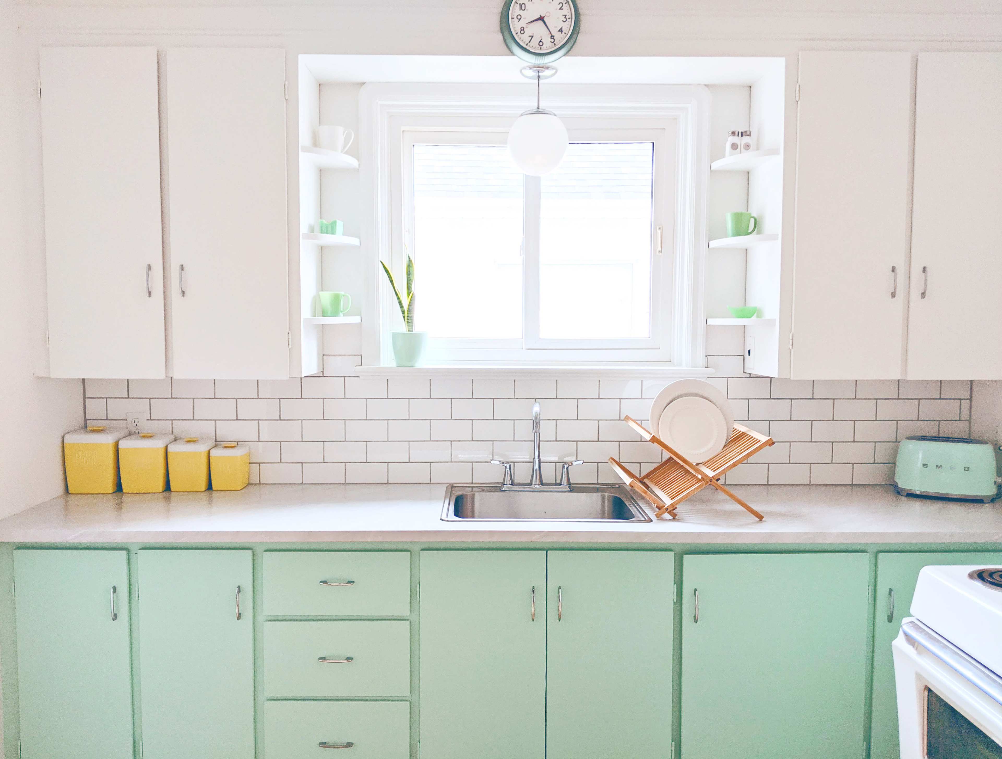 A bright kitchen featuring mint green cabinets, white countertops, and a window over the sink with dishes drying on a wooden rack.