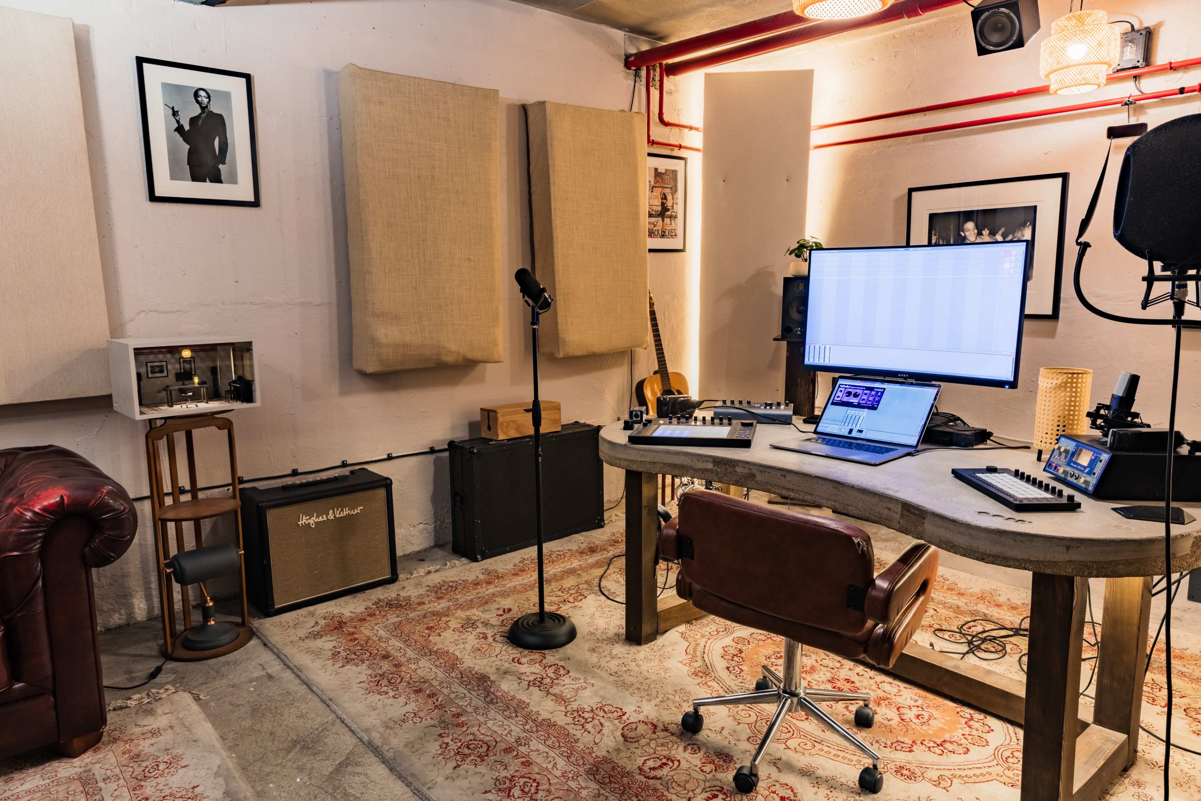 The image shows a music production studio featuring a desk with a laptop, musical equipment, a microphone stand, and sound-absorbing panels on the walls.