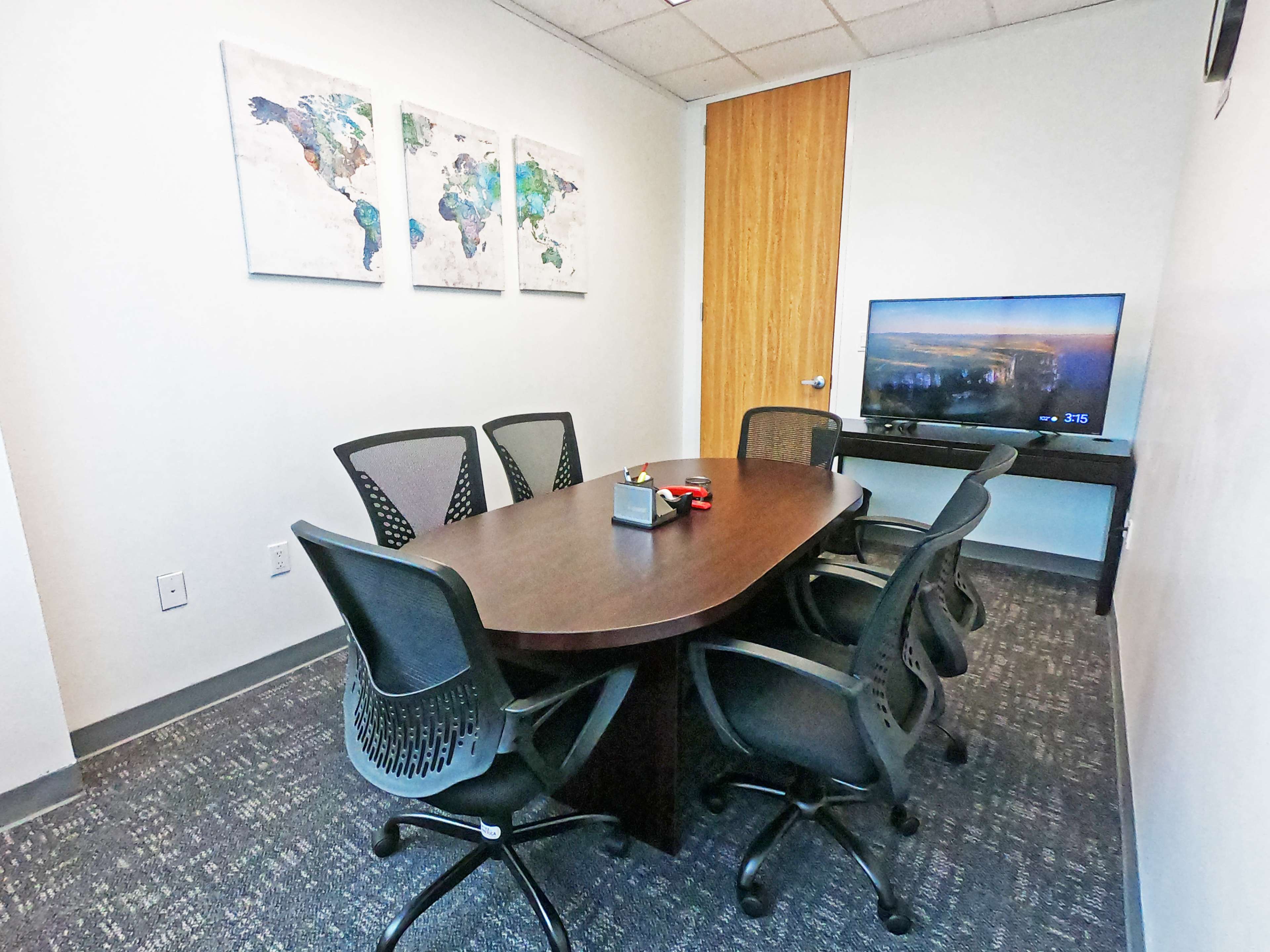 A conference room features a large oval table surrounded by six black mesh chairs, with a television mounted on the wall and three world map art pieces hanging above.