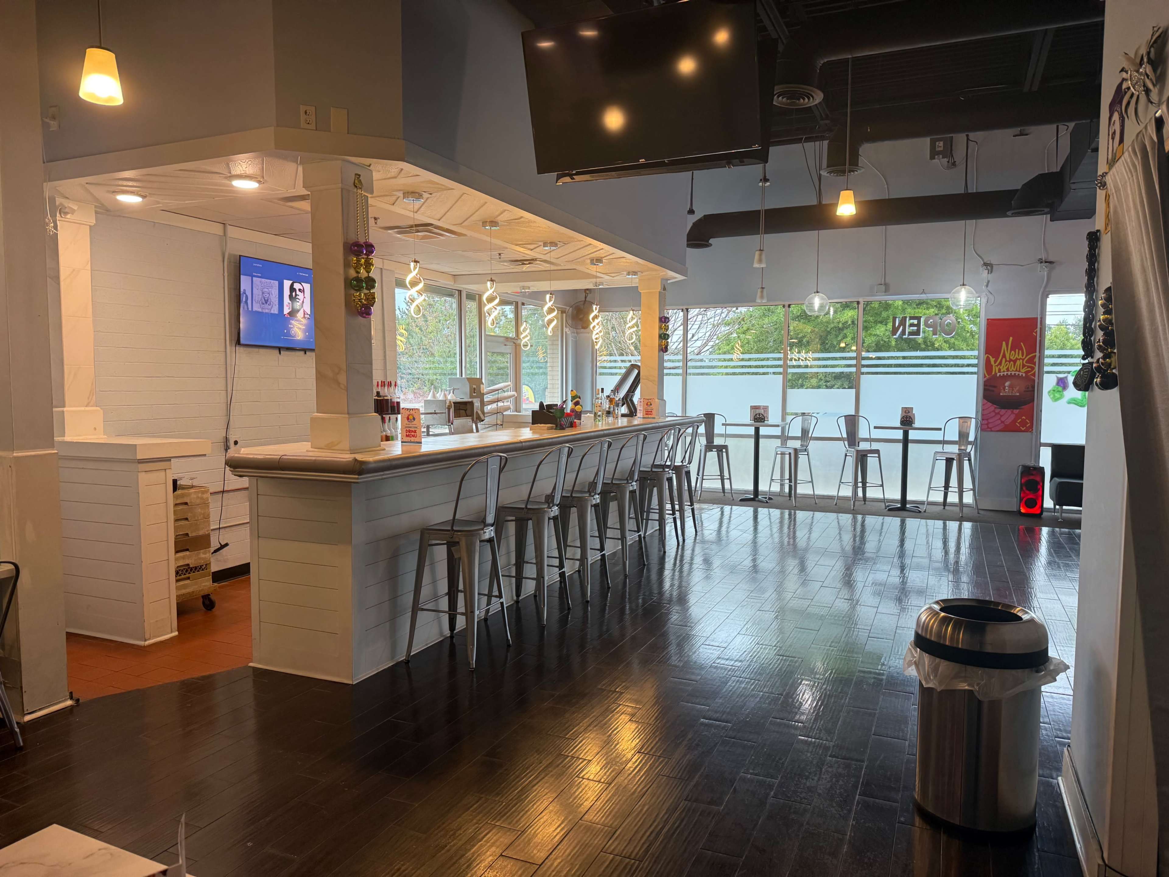 The image shows a restaurant interior featuring a long bar with high stools, a large window overlooking an outdoor area, and a few tables set up in front of the glass.