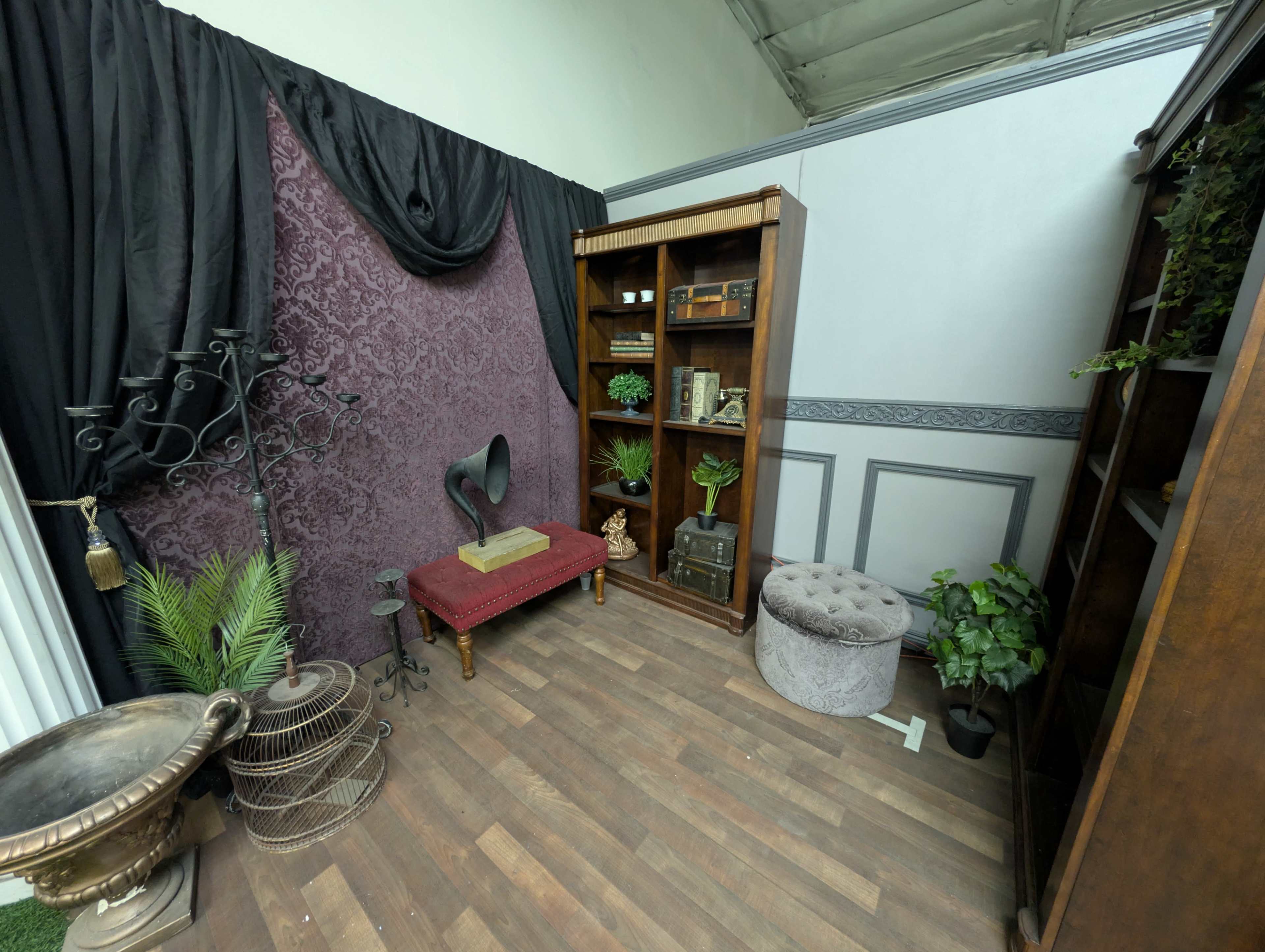 The image shows a vintage-style corner room featuring a red upholstered bench, wooden bookshelves filled with decorative items, a gramophone, and various plants arranged around a wooden floor.